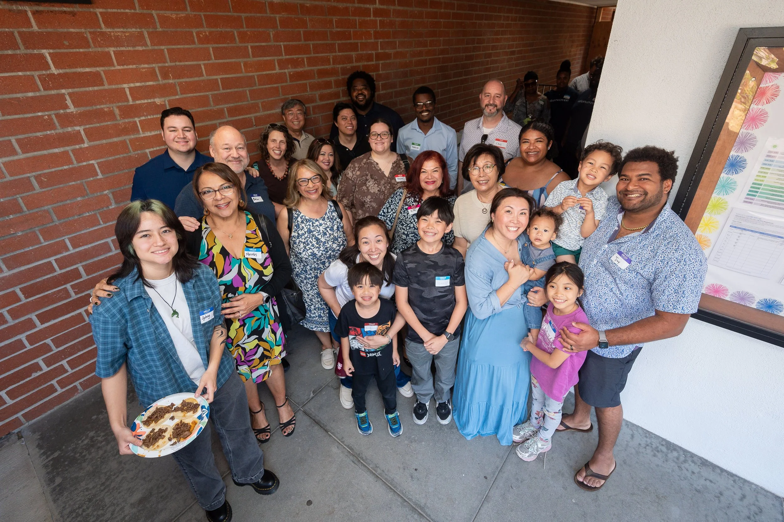 A large group of diverse people including children, adults, and seniors, standing together at a social gathering outdoors near a brick wall, smiling for the photo.