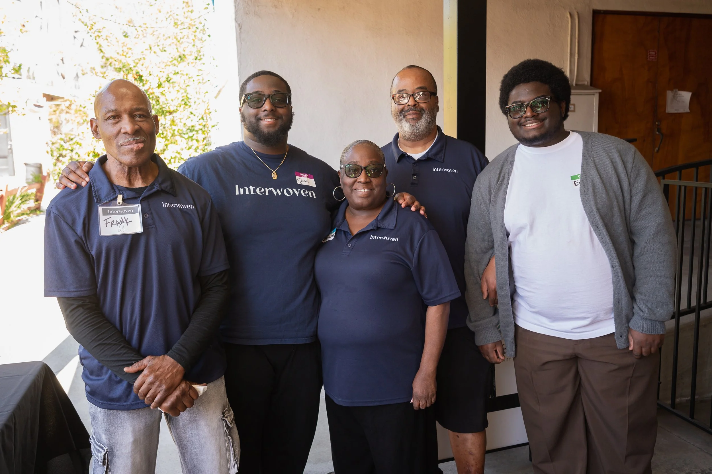 Group of five people posing for a photo, wearing Interwoven branding shirts, standing outside near a building entrance.