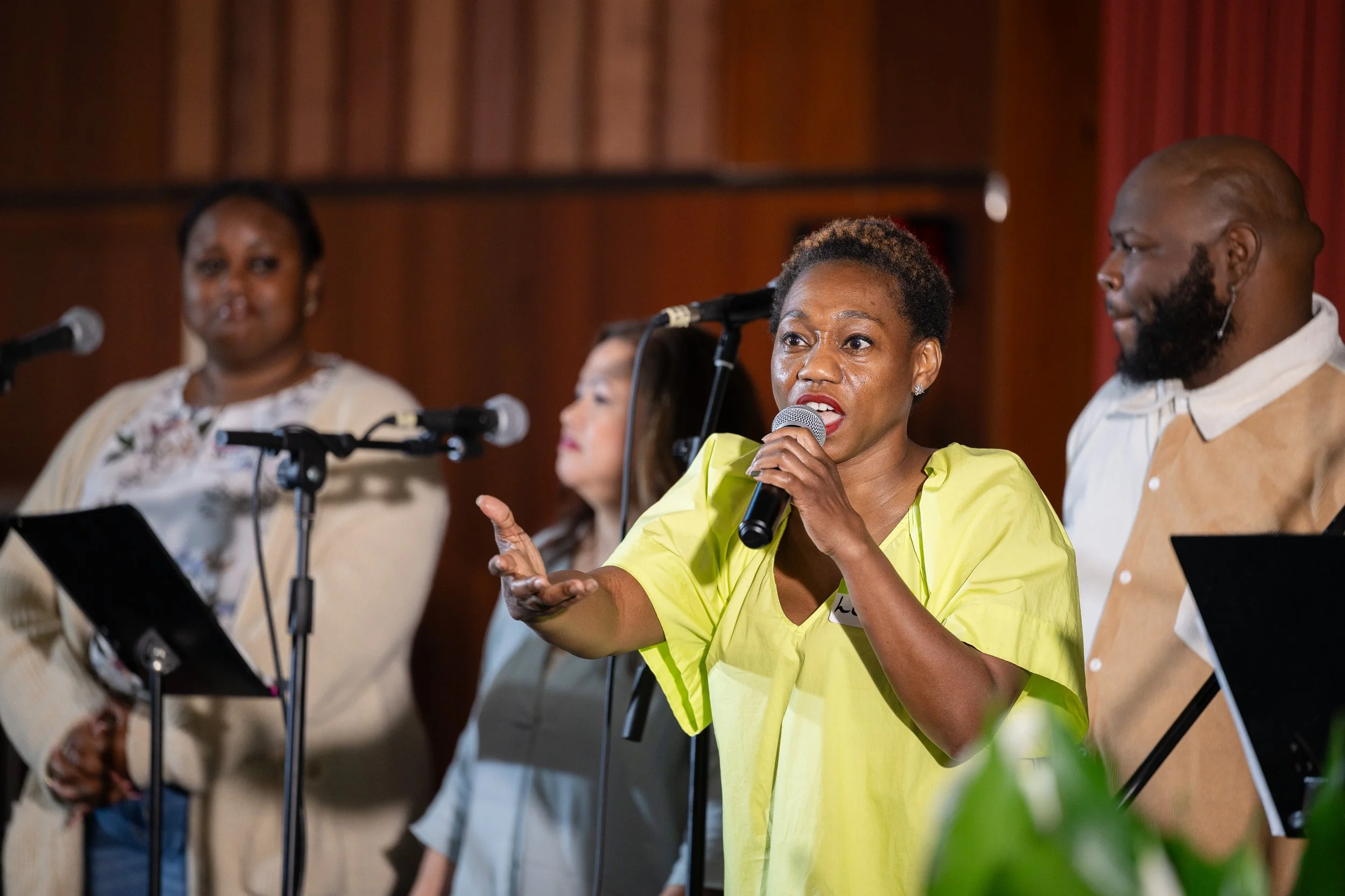 A woman wearing a yellow shirt sining into a microphone on stage, with two women and a man standing behind her, all in front of a wooden-paneled background.