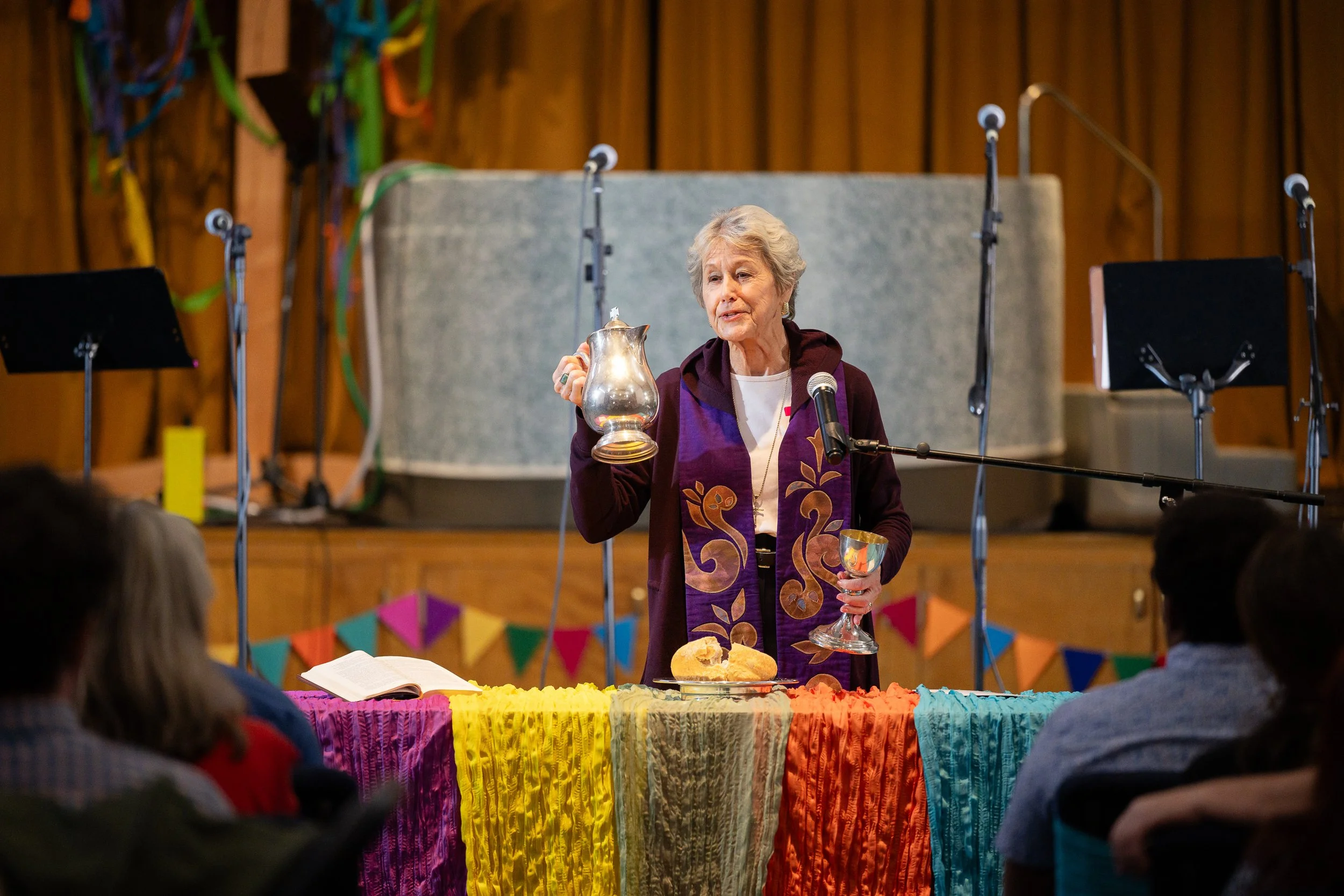 An elderly woman at a celebration or church event holding a silver pitcher in one hand and a goblet in the other, standing behind a table with colorful fabric and food, speaking to a seated audience with microphones on stands in the background.
