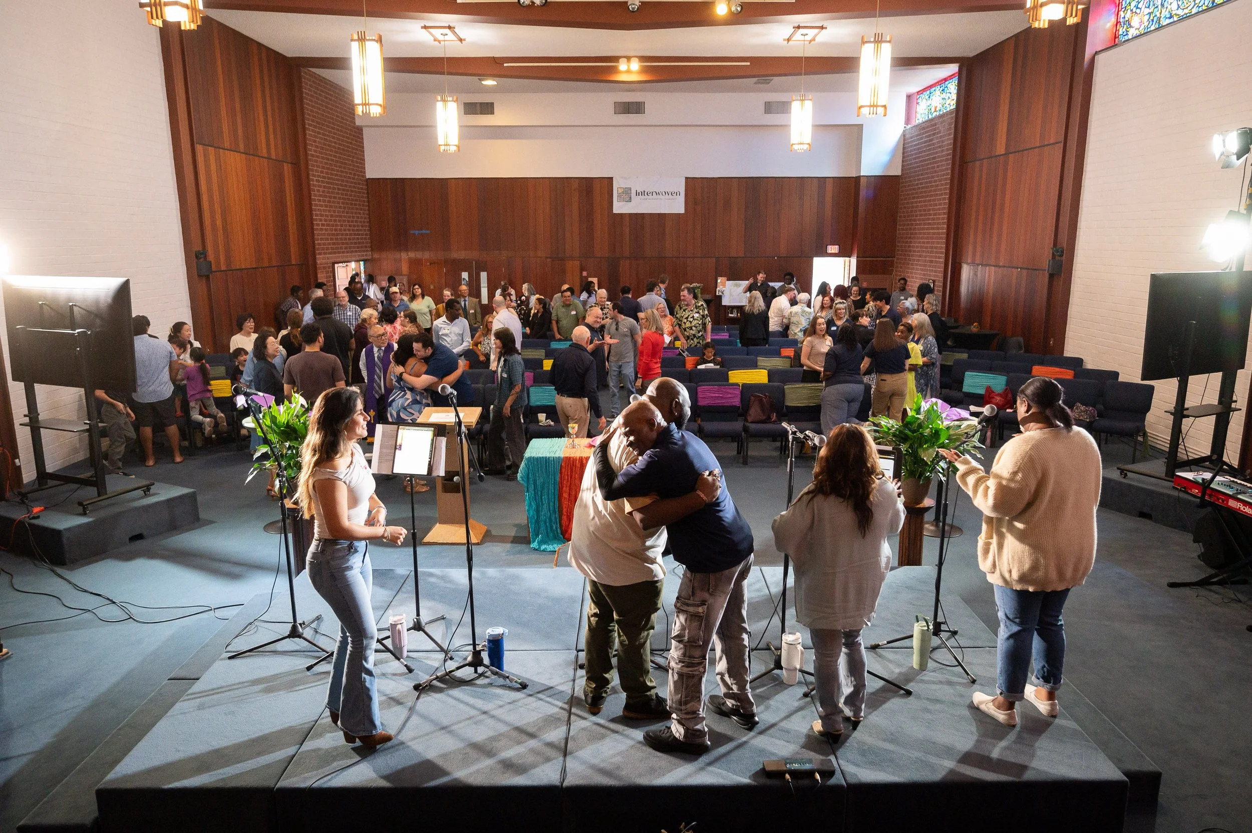 People gathered in a church hall for a Sunday service, with two people hugging in the foreground, and others talking and hugging.