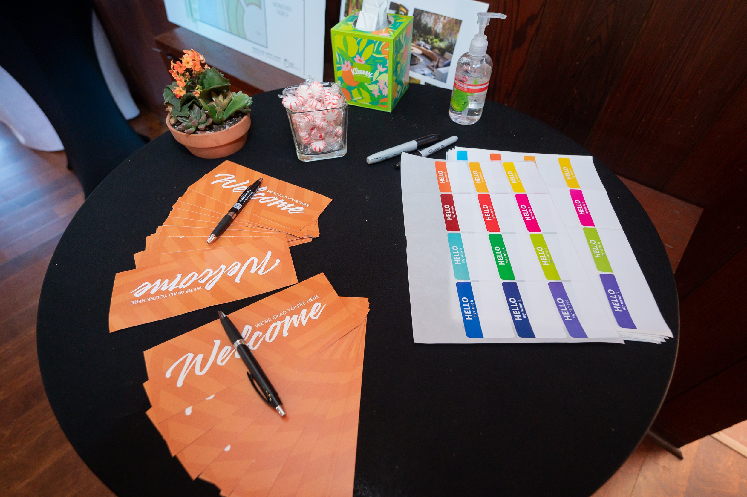 A round black table with orange welcome flyers, black pens, a stack of colorful name tags, a small potted succulent plant, a glass jar filled with peppermint candies, a box of Kleenex tissues, and a bottle of hand sanitizer, set against a wooden wall background.