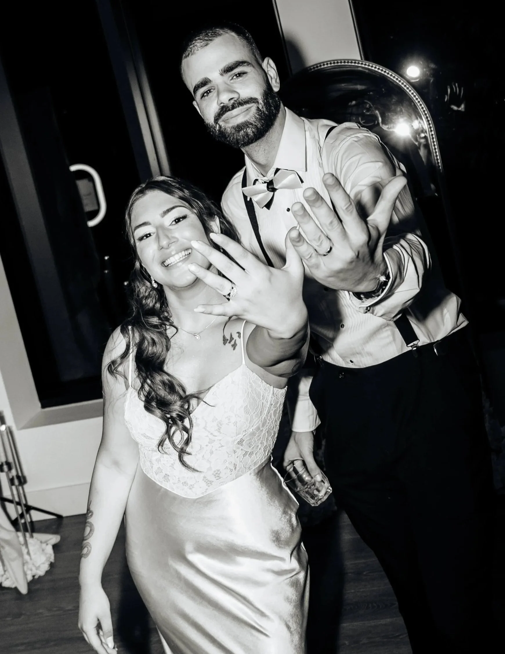 En noir et blanc, une femme et un homme tendent le bras, tenant chacun une bague de mariage dans leurs doigts de la main gauche
