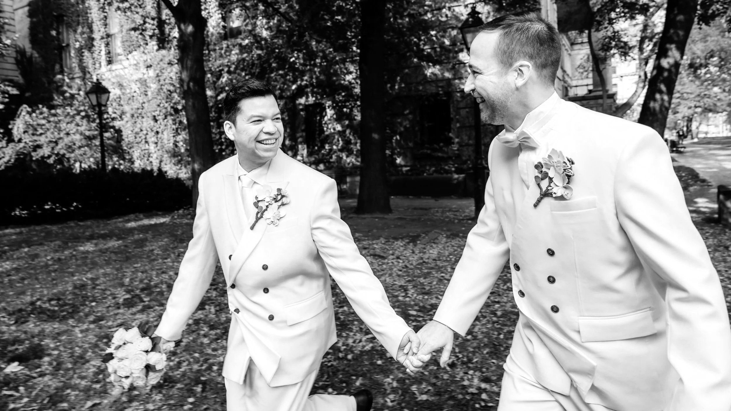 Deux hommes en habit de mariés courent dans des feuilles d’automne, l’un d’eux tenant un bouquet de fleurs jaunes