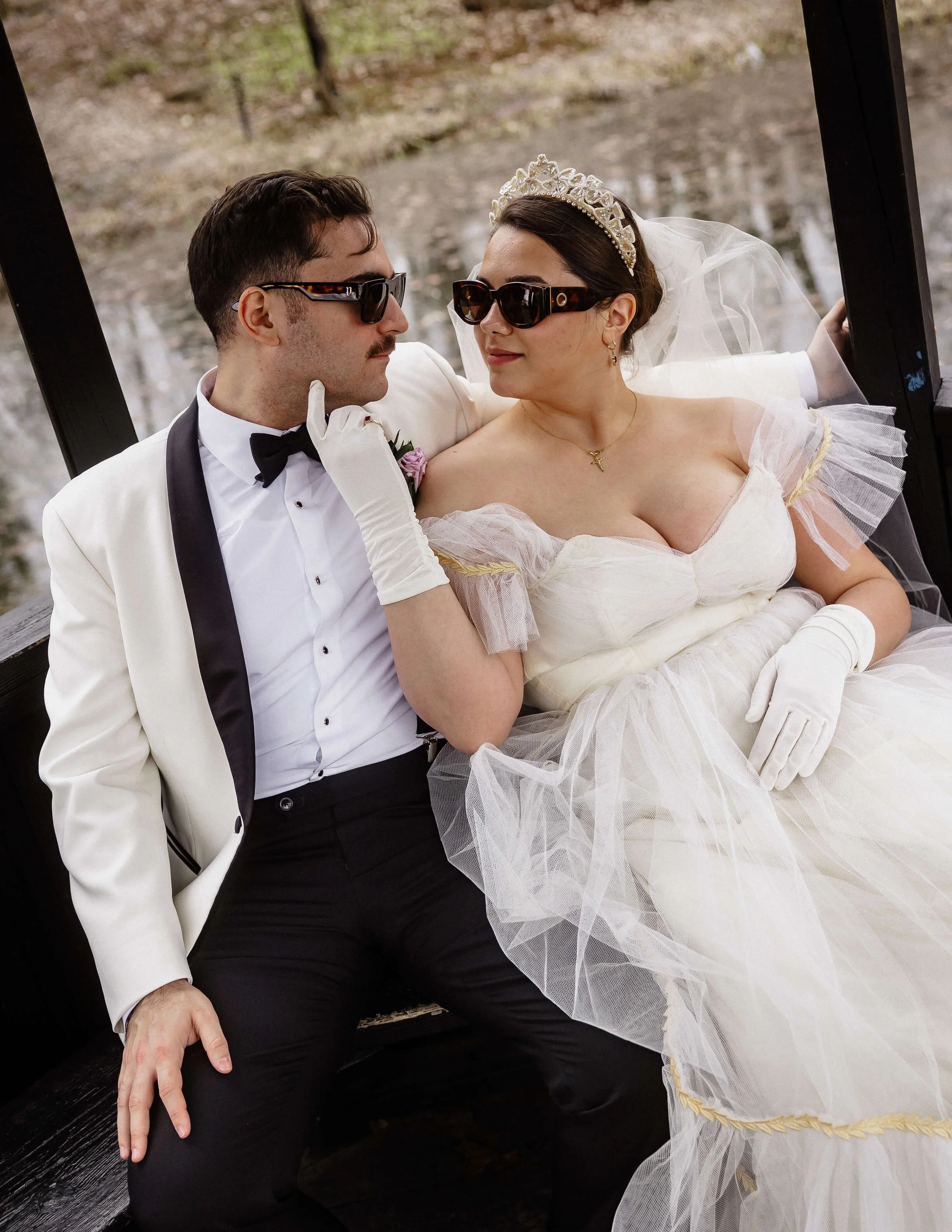 Sur un banc dans un parc, un homme et une femme se regardent. Ils portent des lunettes de soleil et des habits de mariage.