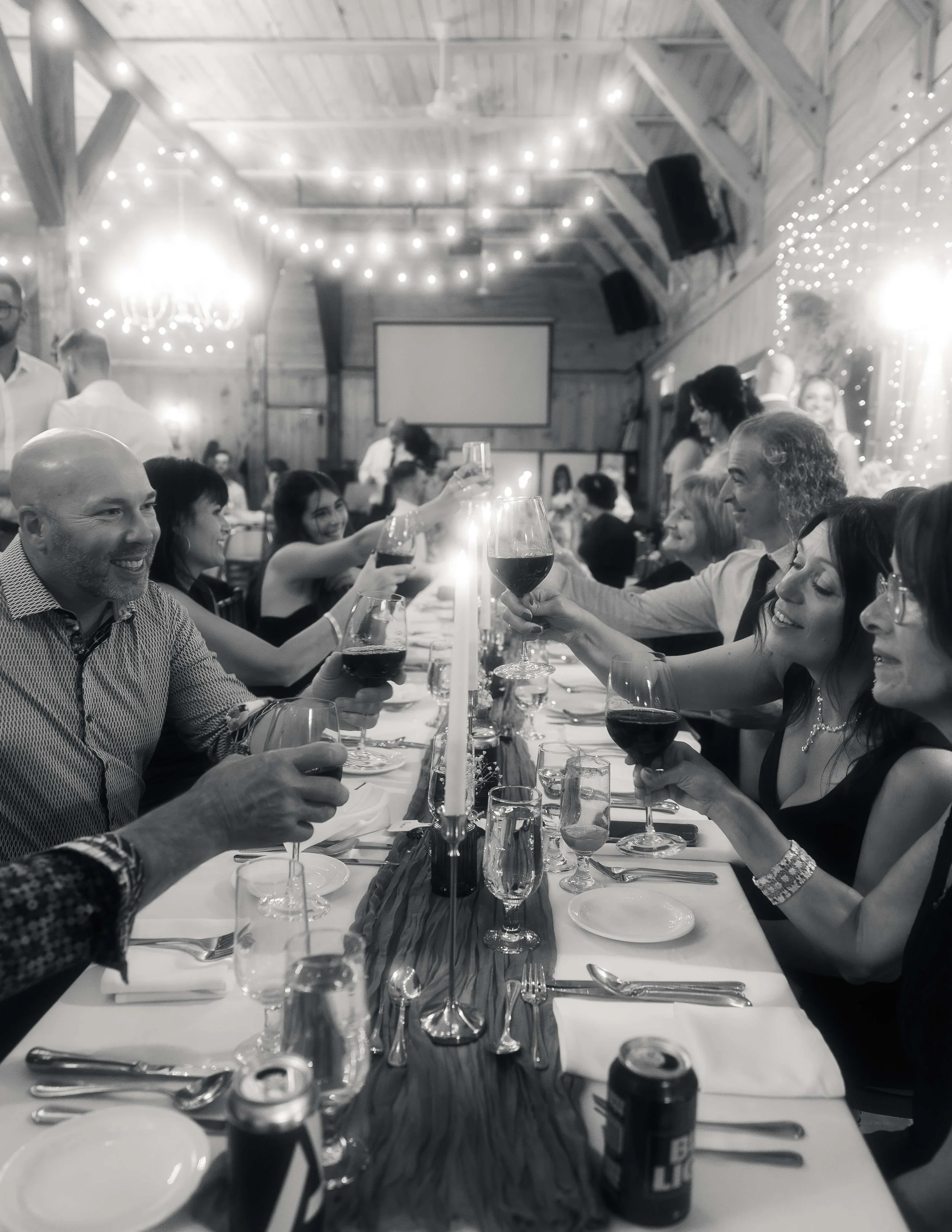 Groupe de personnes à une célébration de mariage assis autour d'une longue table, levant leurs verres pour faire un toast aux mariés, dans une salle décorée avec des guirlandes lumineuses