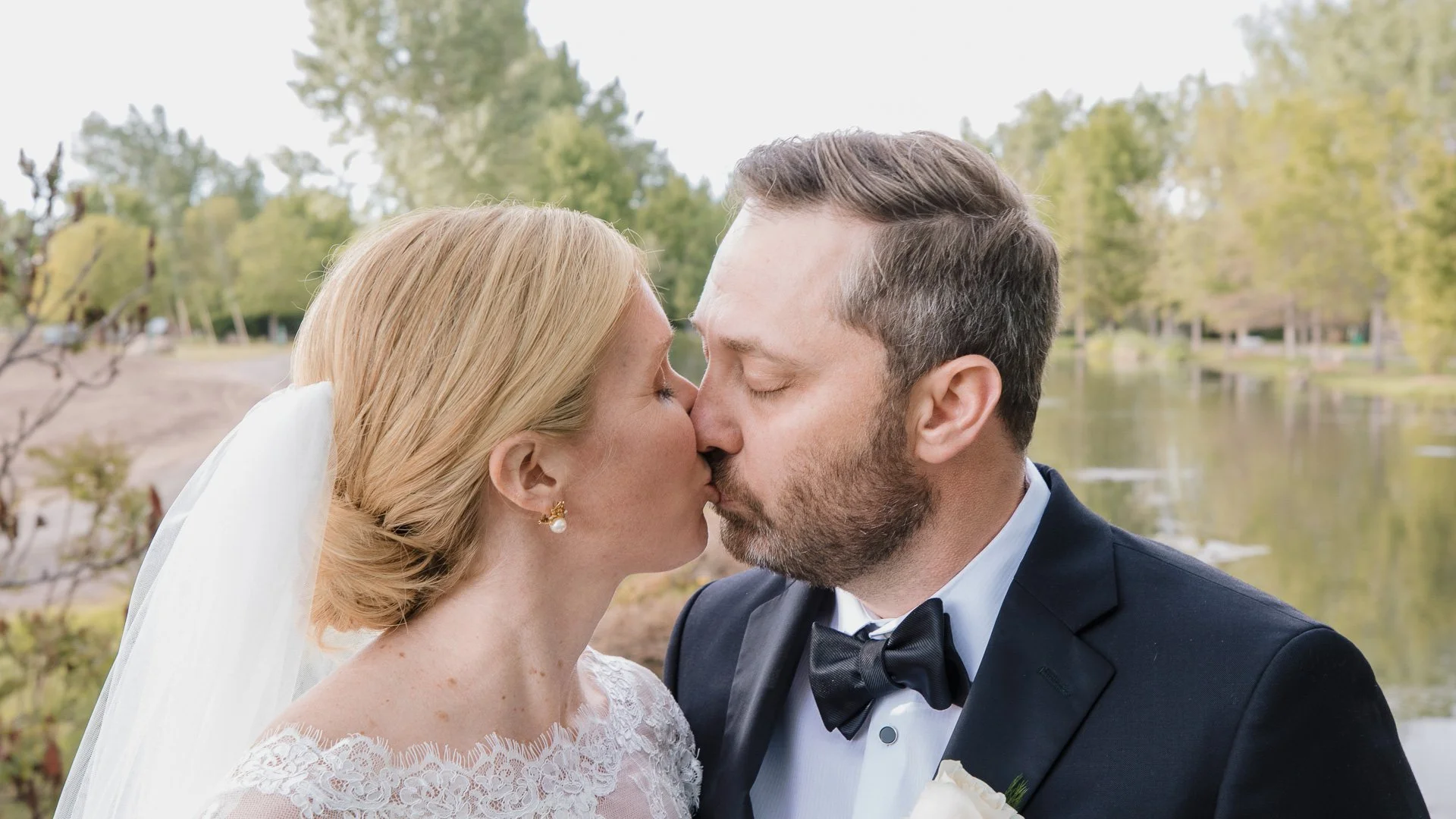 A bride and groom kissing outdoors near a lake with trees in the background.