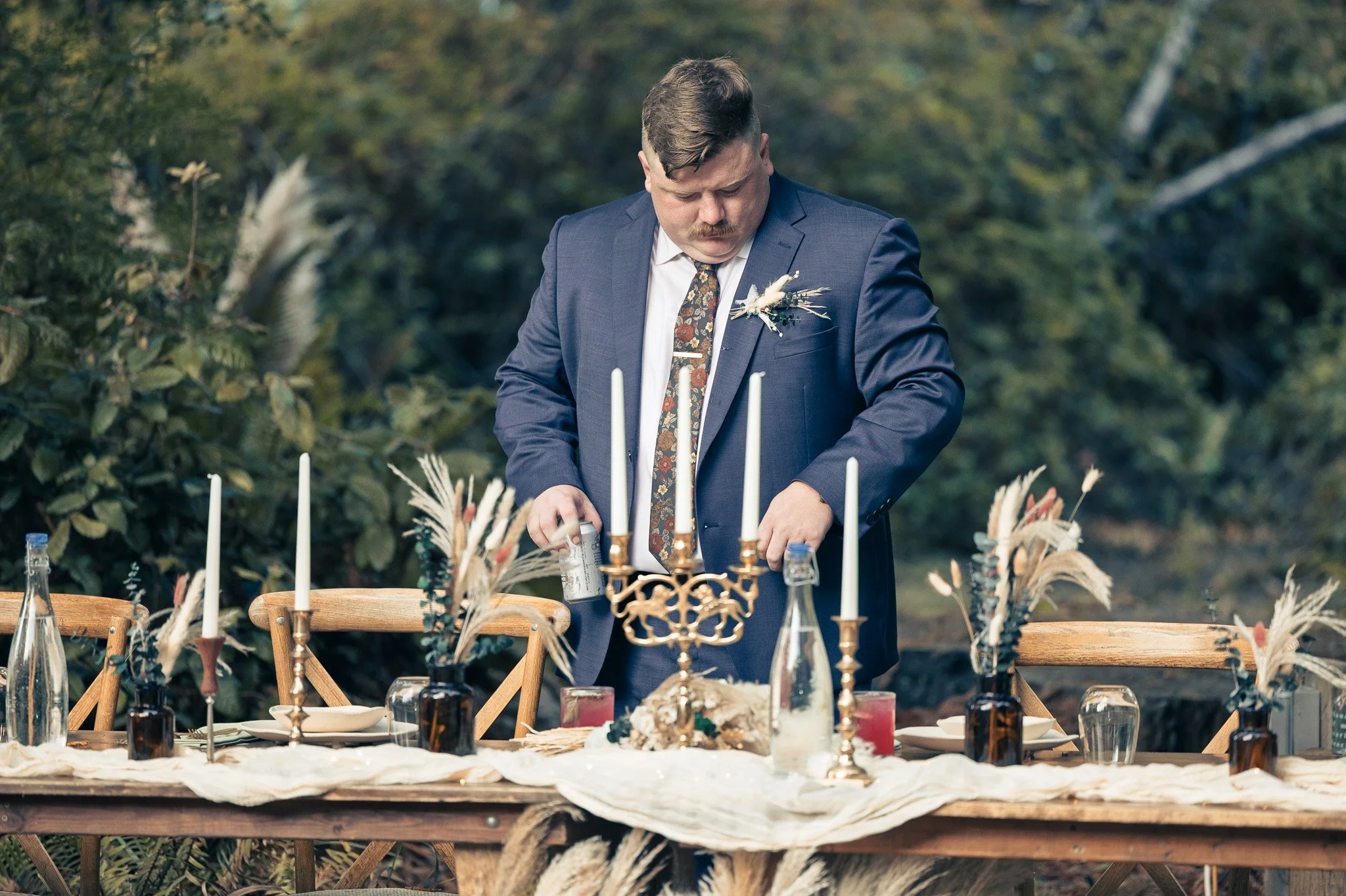 Man in navy suit with floral tie and boutonniere standing at outdoor wedding reception table, adjusting items on the table with a lush forested background.