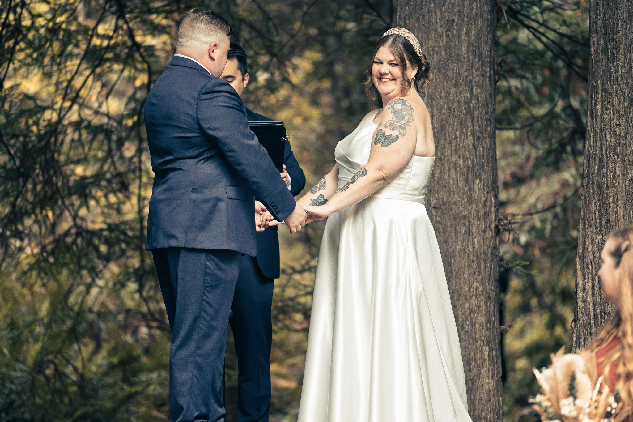 A woman in a white wedding dress holding hands with a man in a blue suit during a wedding ceremony outdoors among trees.