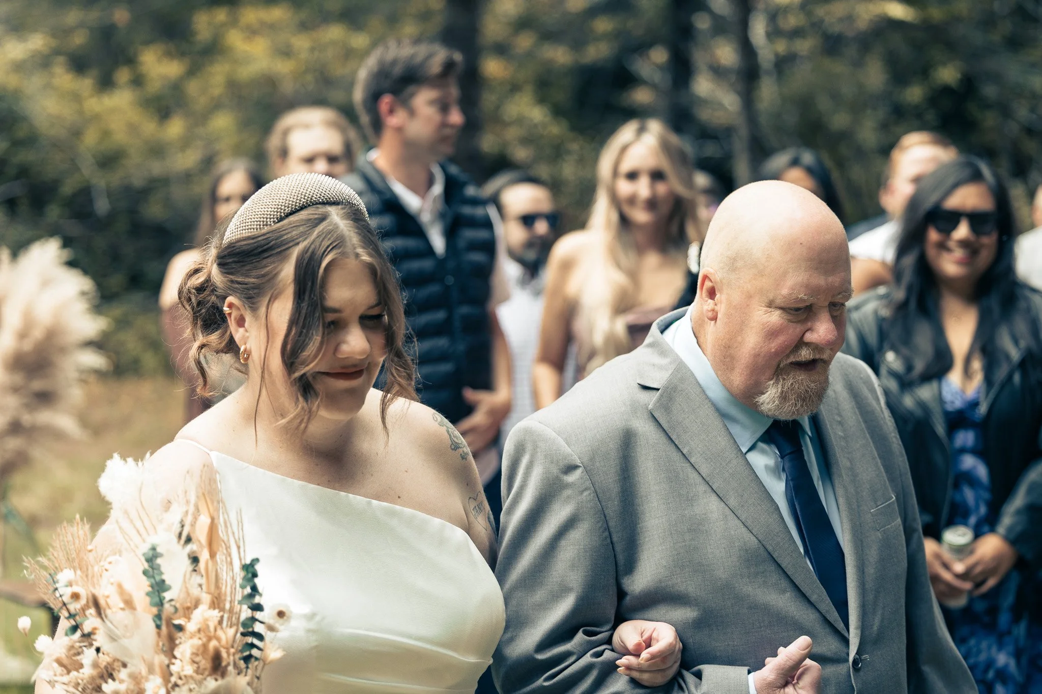 A young woman in a white dress with a pearl headband and a bouquet of dried flowers is walking arm-in-arm with an older man in a gray suit during an outdoor event, surrounded by a group of smiling people.