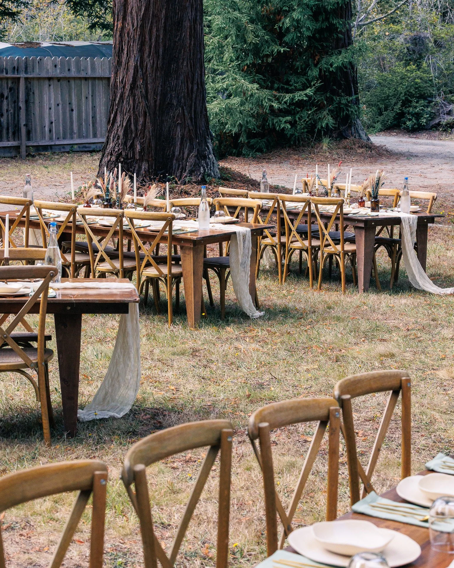 Outdoor dining setup with wooden tables and chairs, decorated with bottles, candles, and natural centerpieces, set among large trees in a forested area.