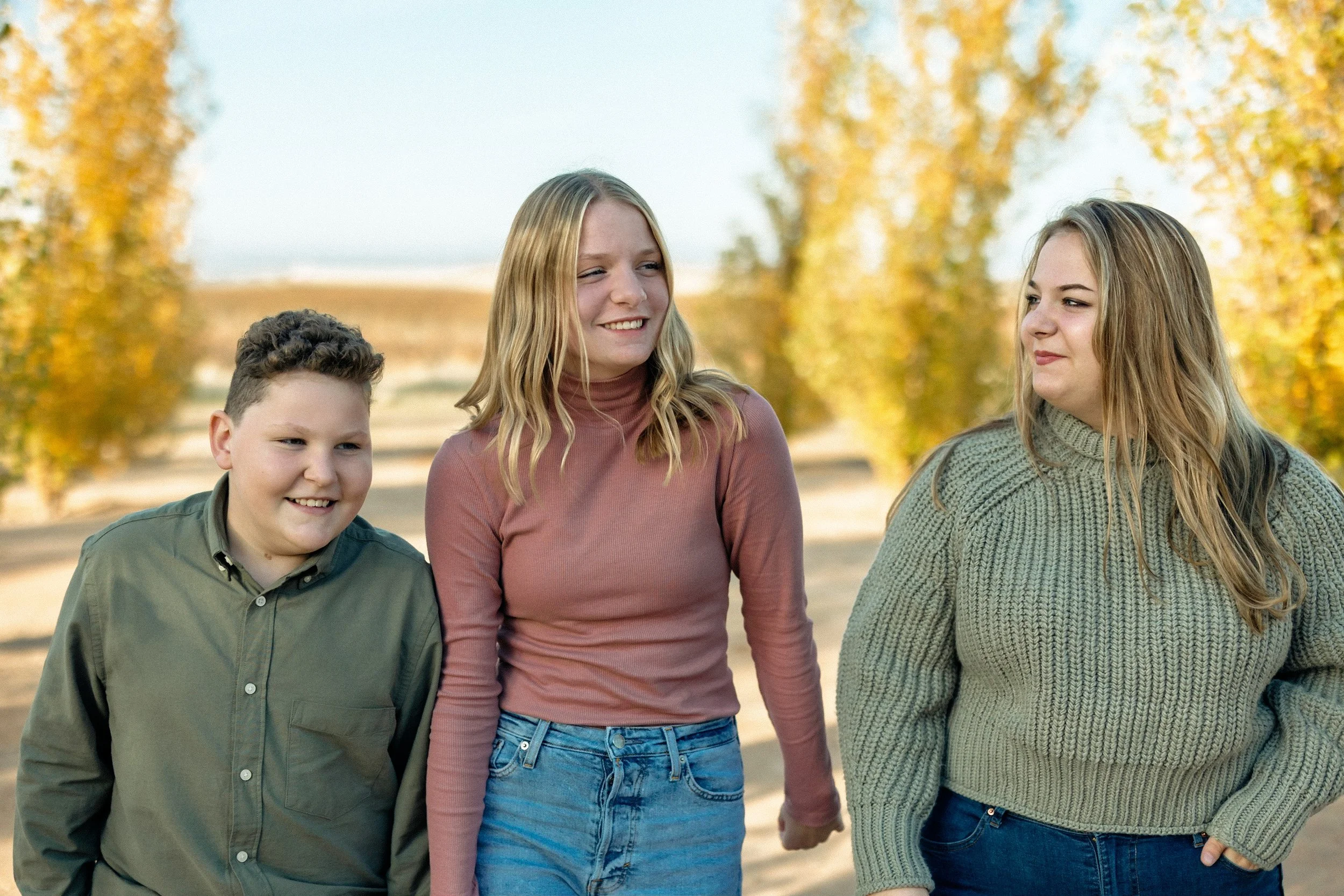 Three teenagers, two girls and one boy, walking outdoors on a sunny day during autumn, with yellow-leaved trees in the background. The girl in the middle is smiling and wearing a pink long-sleeve shirt and blue jeans. The girl on the right is looking at her and wearing a green sweater. The boy on the left is smiling and wearing a green button-up shirt.