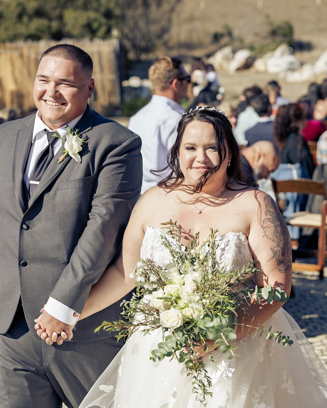 Bride and groom holding hands during outdoor wedding ceremony, with guests in the background.