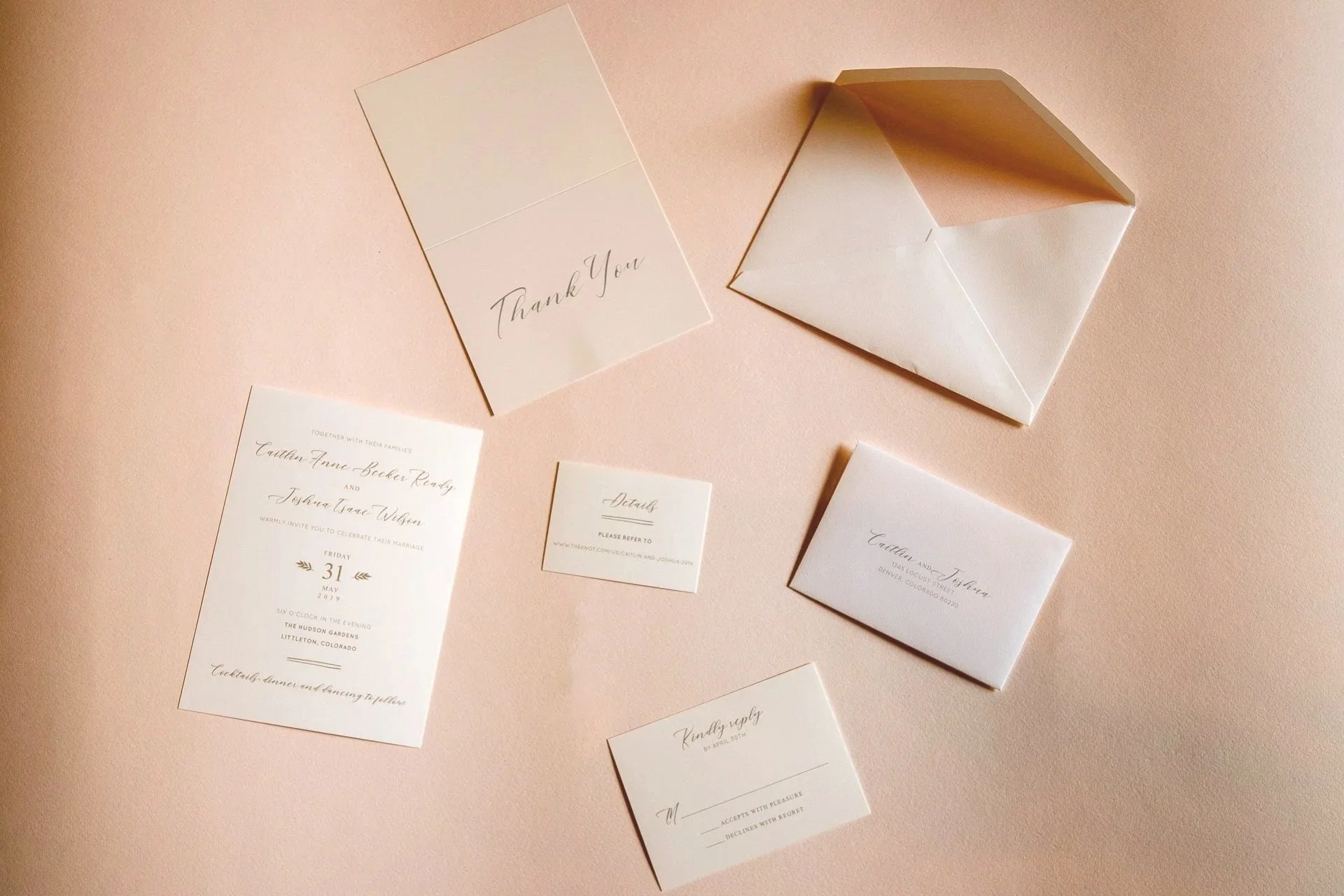 A collection of wedding invitation stationery including a thank you card, an envelope, an invitation, response cards, and details cards, all arranged on a pink surface.