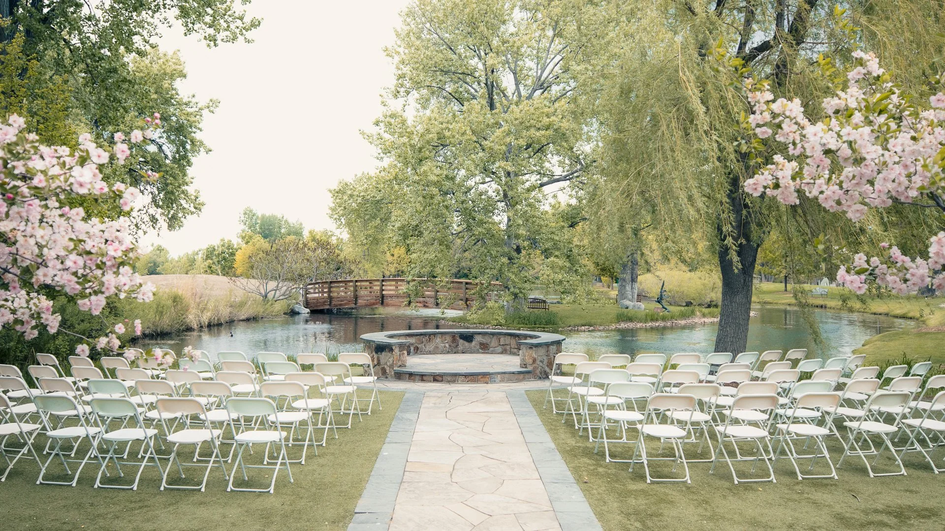 Outdoor wedding ceremony setup with rows of white chairs on a grassy area facing a small stage with a water backdrop, surrounded by trees and flowering bushes.