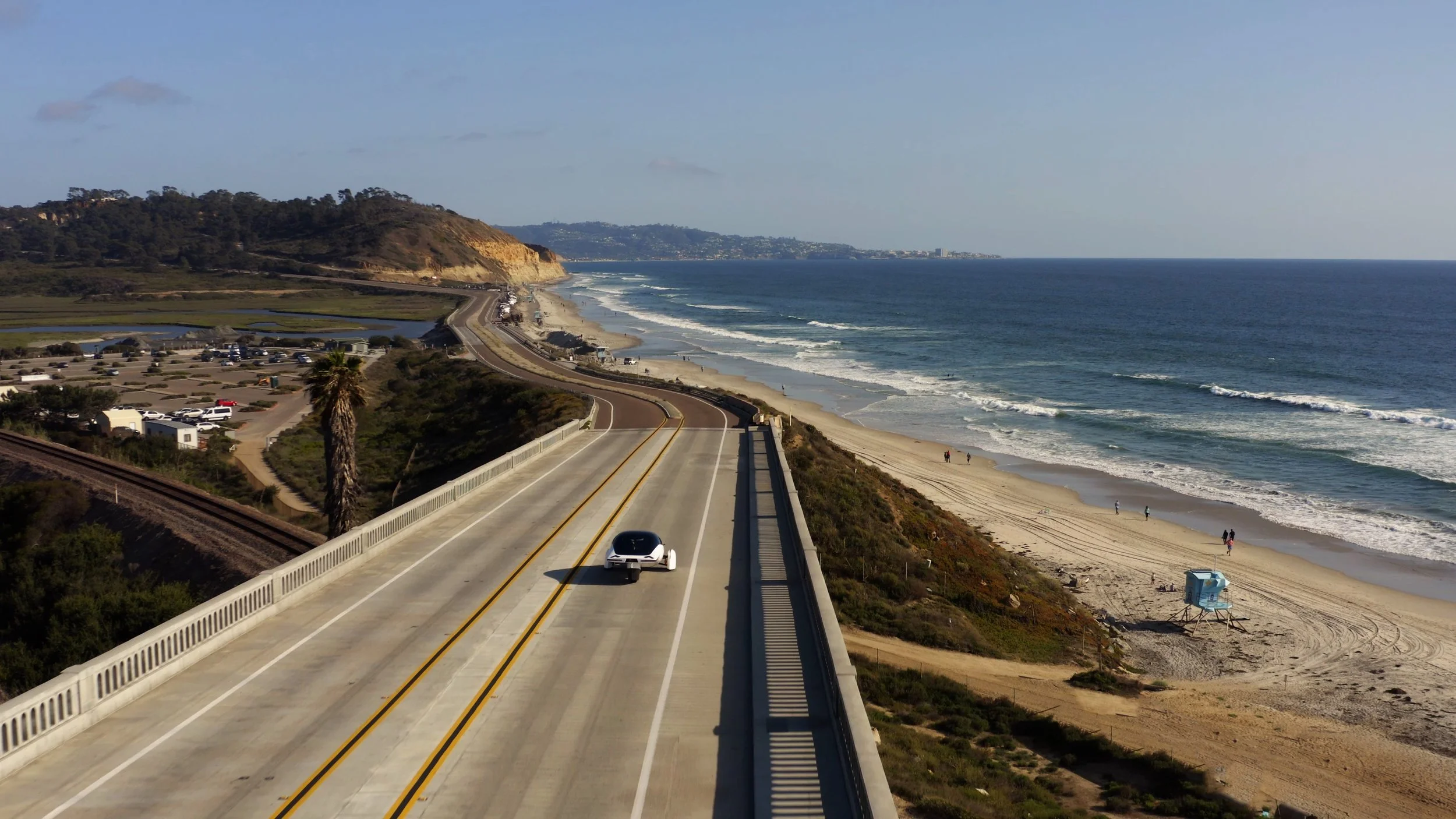 Lifestyle shot of the Aptera vehicle driving over a bridge with the California coast off to the right