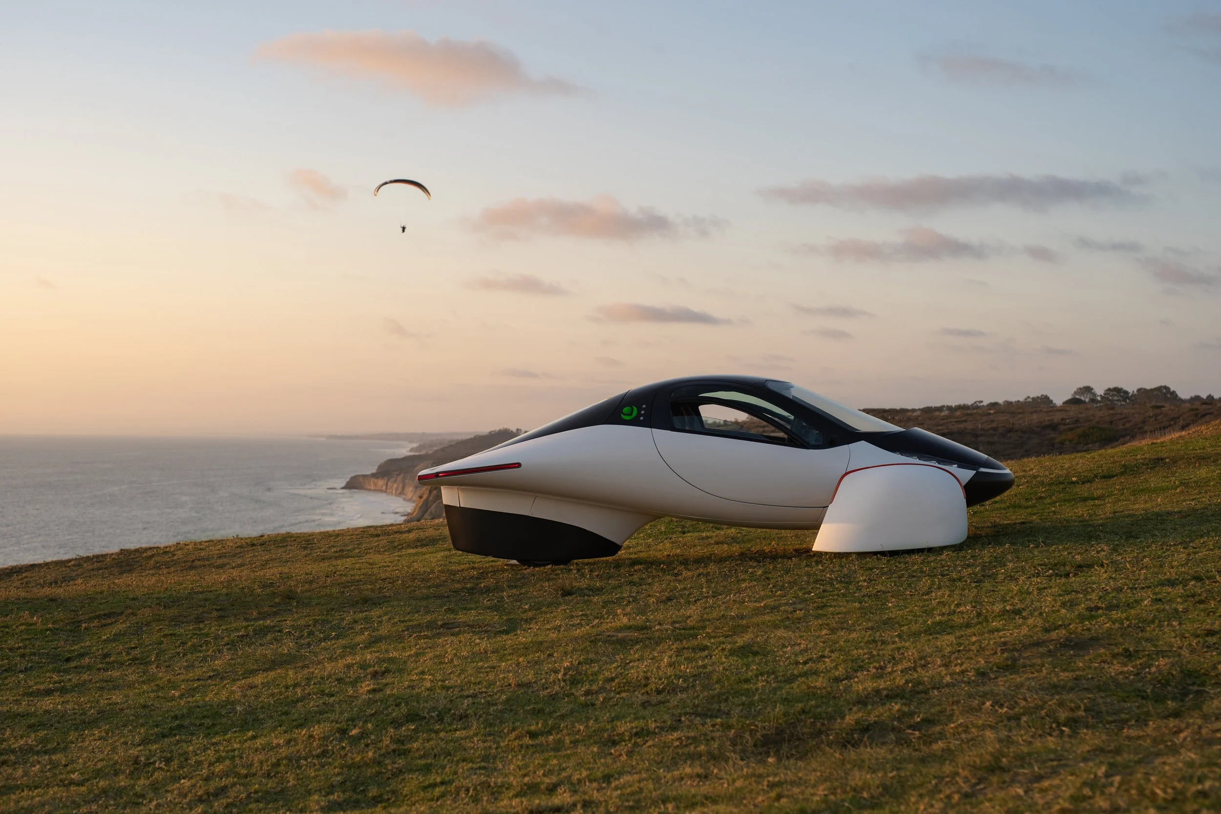 Scenic lifestyle shot of the Aptera vehicle at sunset parked on a high up lookout point with a paraglider off in the distance flying in the sky