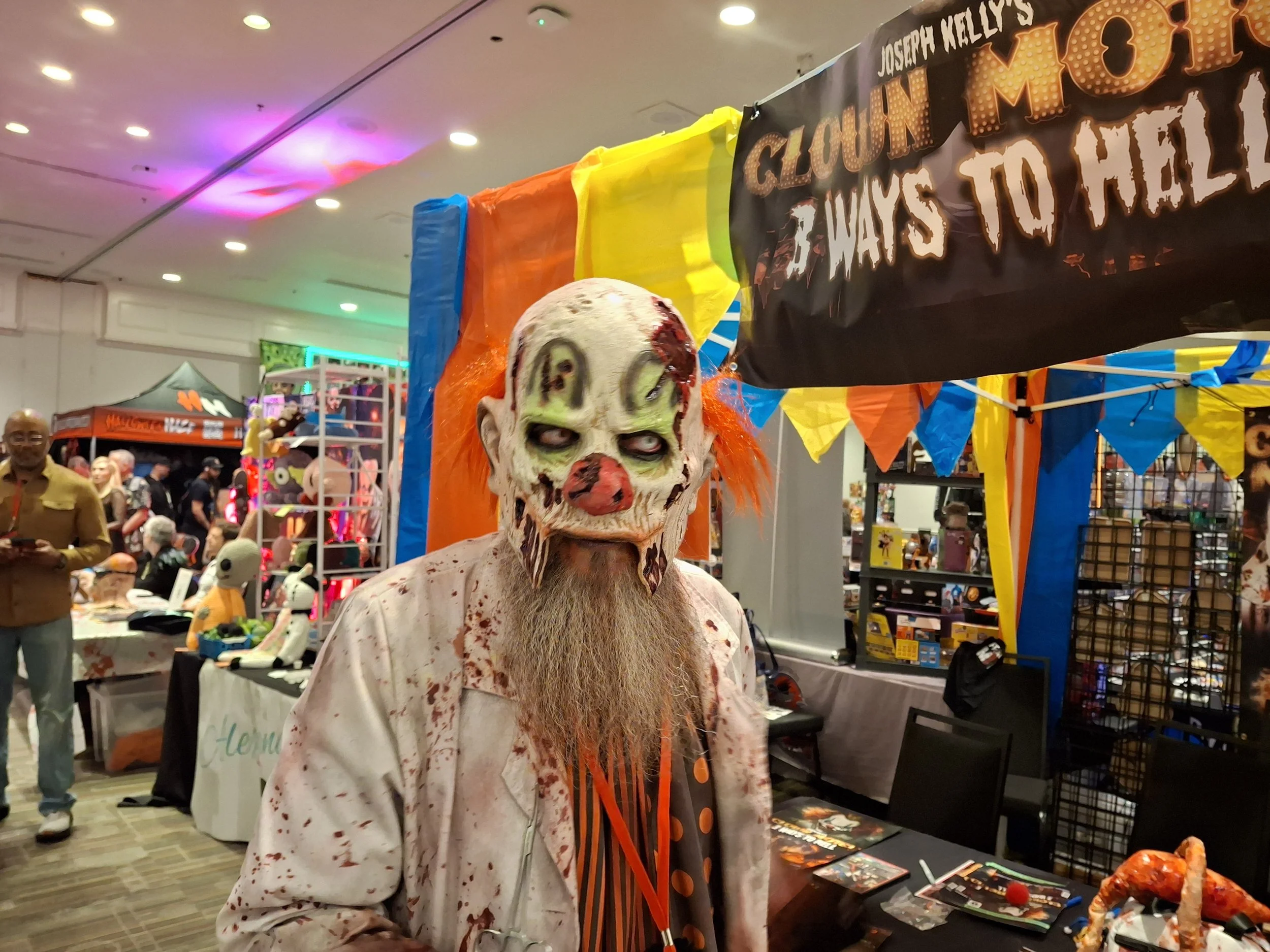 A person dressed as a creepy clown with a bloody white face, green eyes, orange hair, and a long beard at a Halloween or horror-themed event.