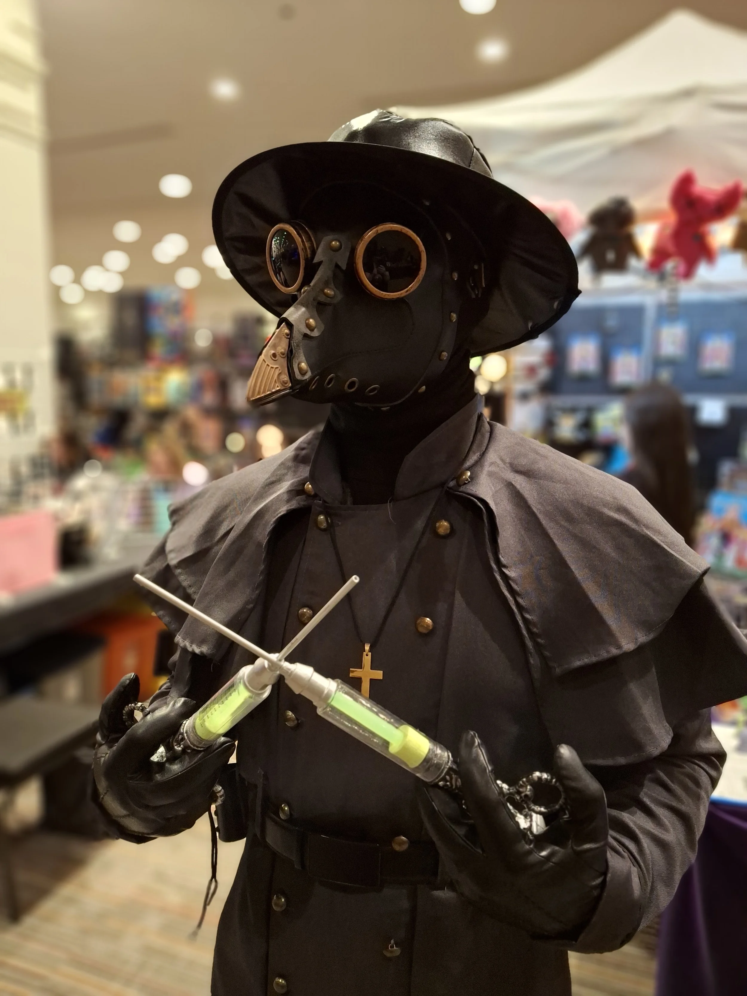 Person dressed as a steampunk villain with a black hat, mask, cape, and gloves holding two syringes with glowing liquids at an indoor event.