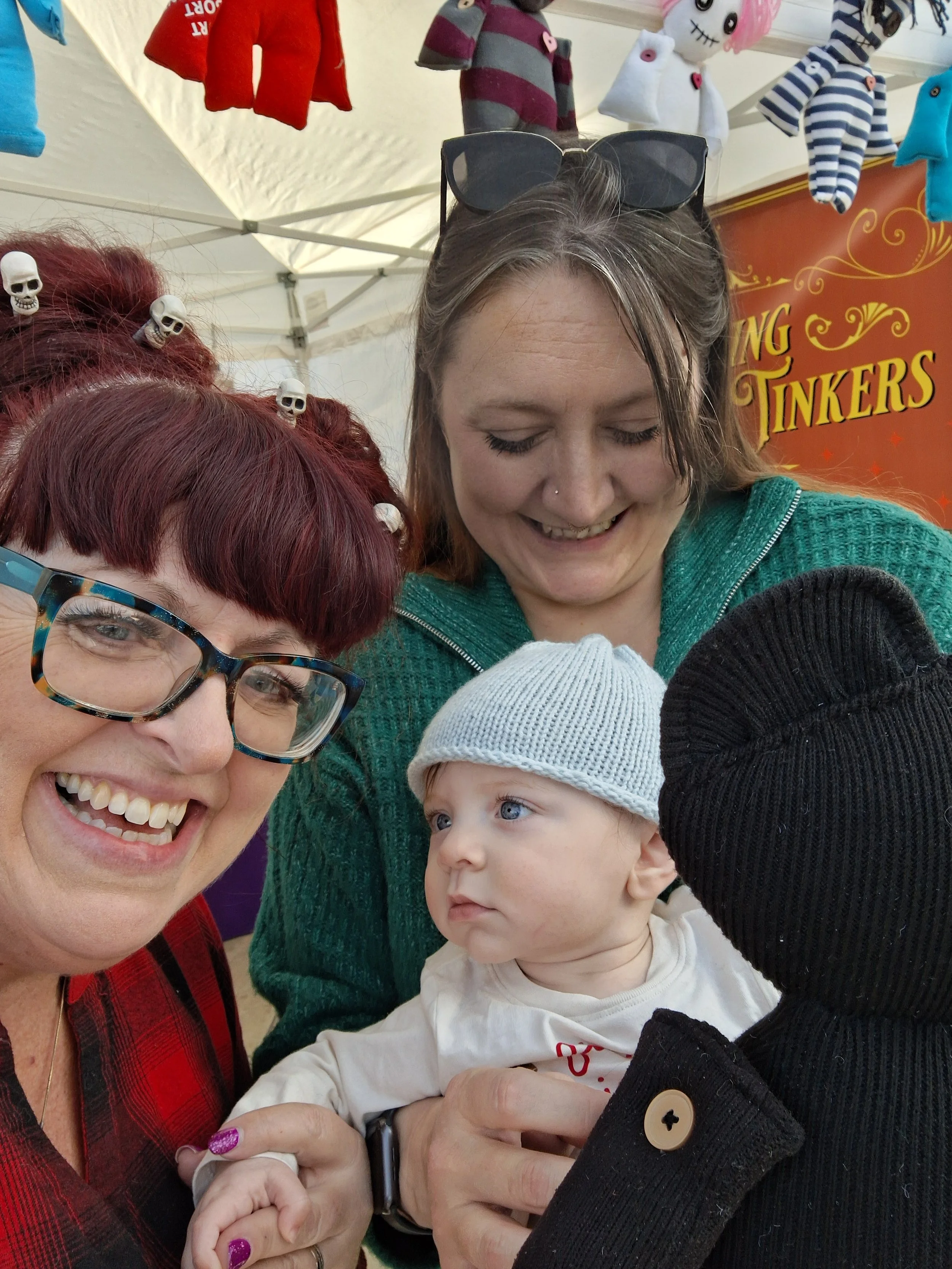 Close-up of two women and a baby at a Halloween-themed event. The woman on the left has short red hair with skull hairpins, glasses, and a red plaid shirt. The woman in the center has shoulder-length brown hair, sunglasses on her head, and a green sw