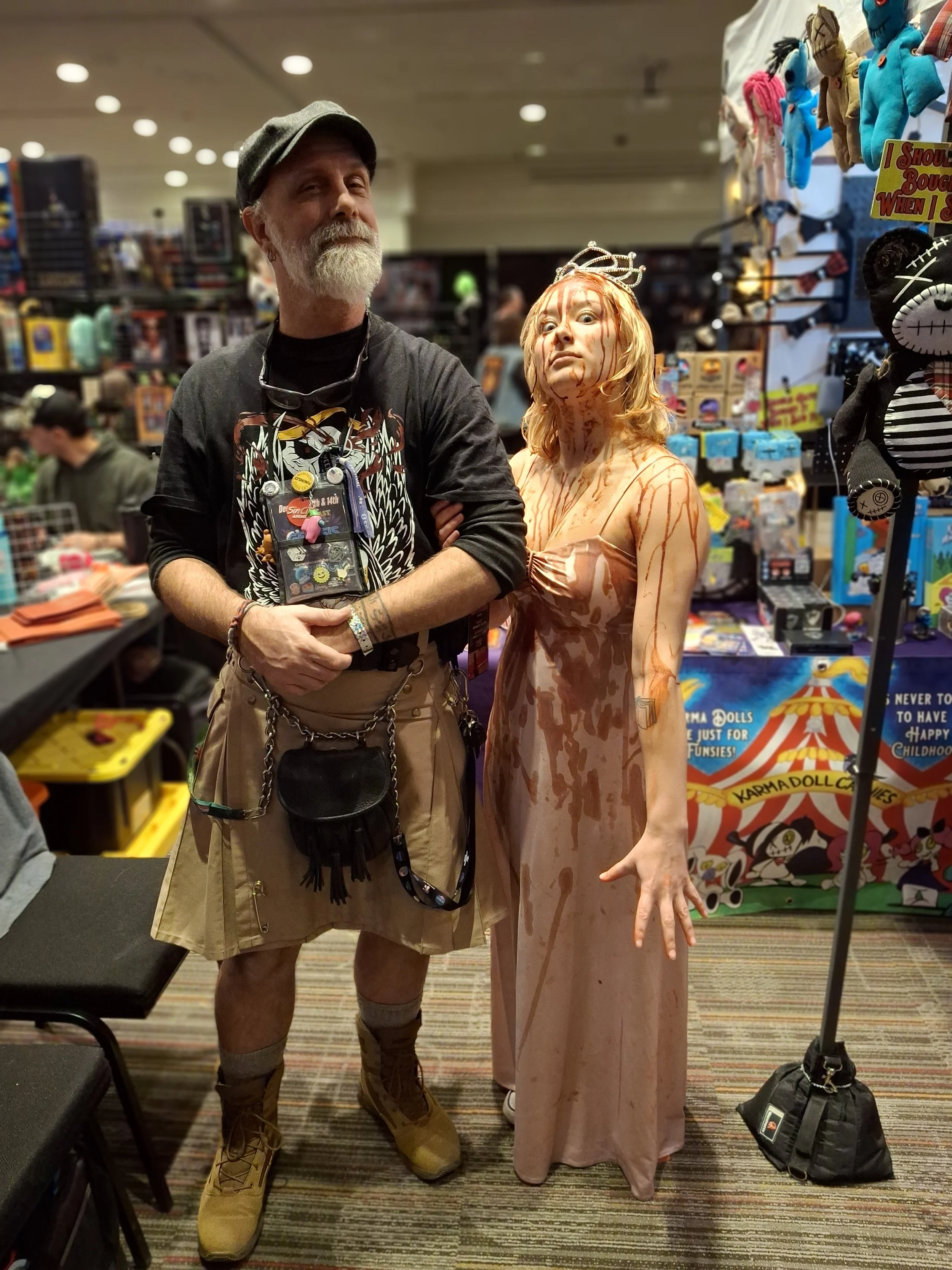 A man with a gray beard and a woman dressed as a bloody zombie in a convention or toy store with shelves of toys and plushies in the background.