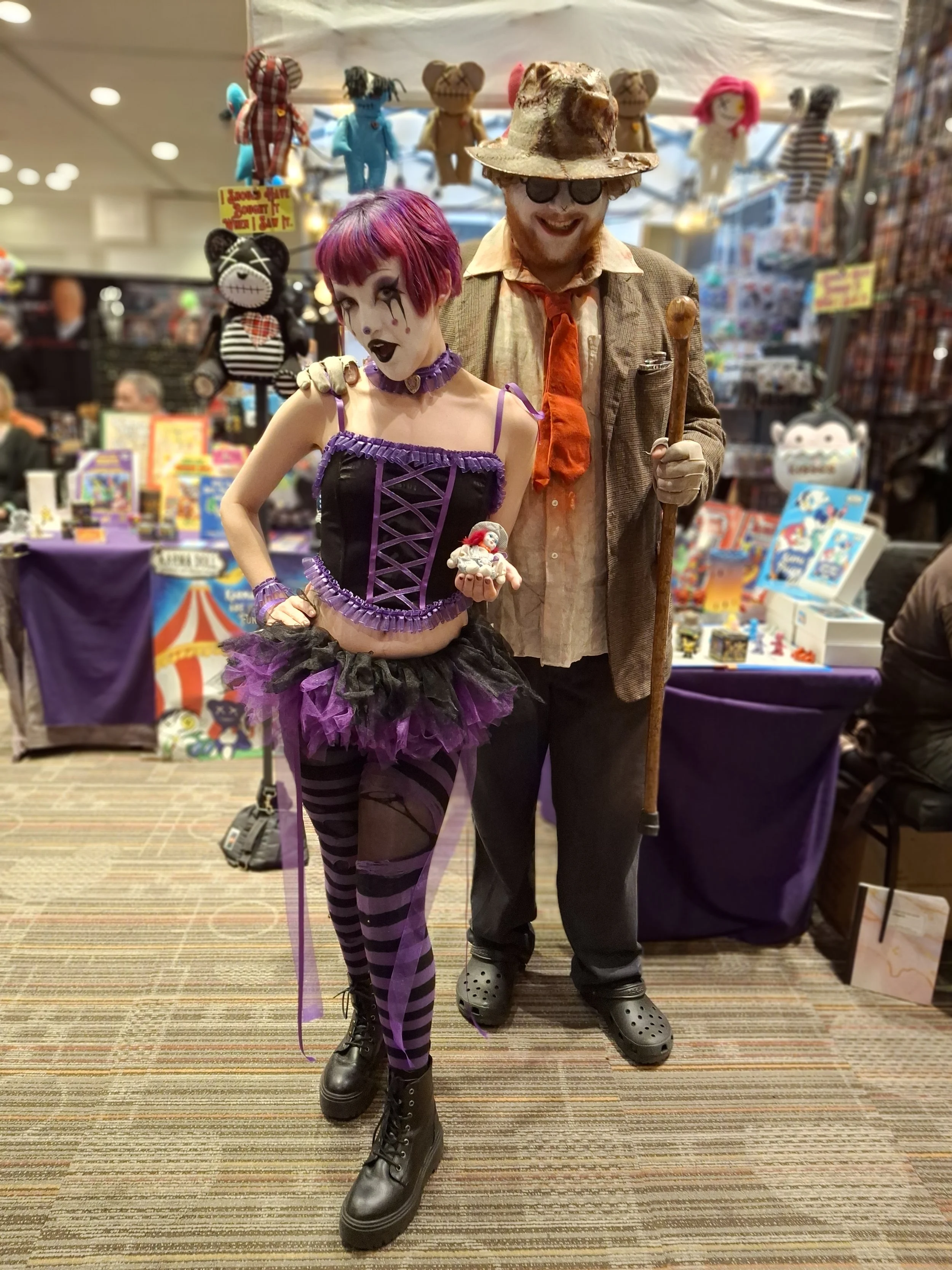 A woman dressed as a goth clown with purple and black makeup, short purple hair, a purple and black corset, and striped leggings, holding a small clown figurine. Beside her, a man dressed as an eccentric old man with a wide-brimmed hat, glasses, an o