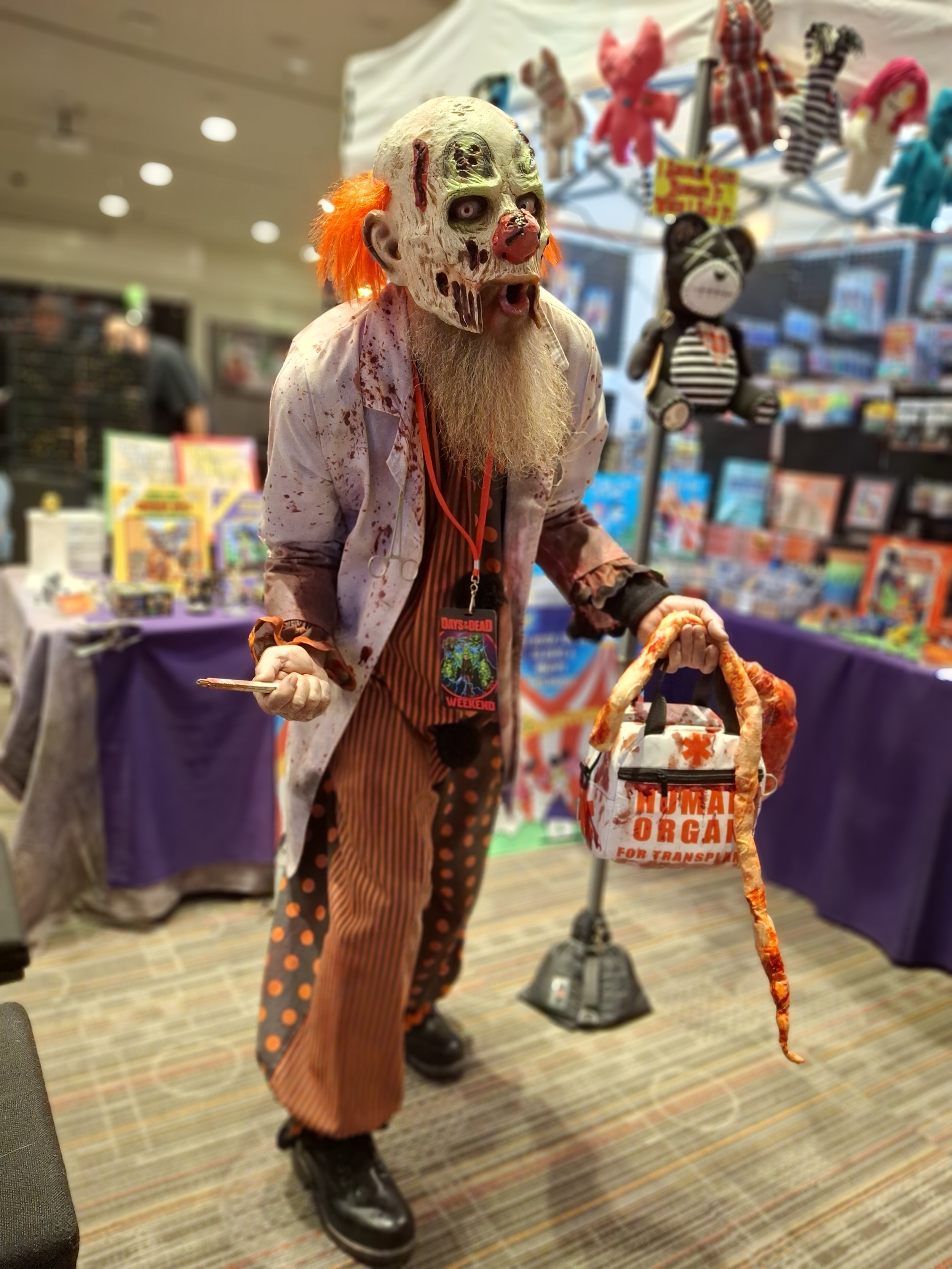 Person dressed as a creepy clown with a mask, orange hair, and blood-stained clothes, holding a fake pizza slice and a money bag, at a Halloween or horror-themed event.