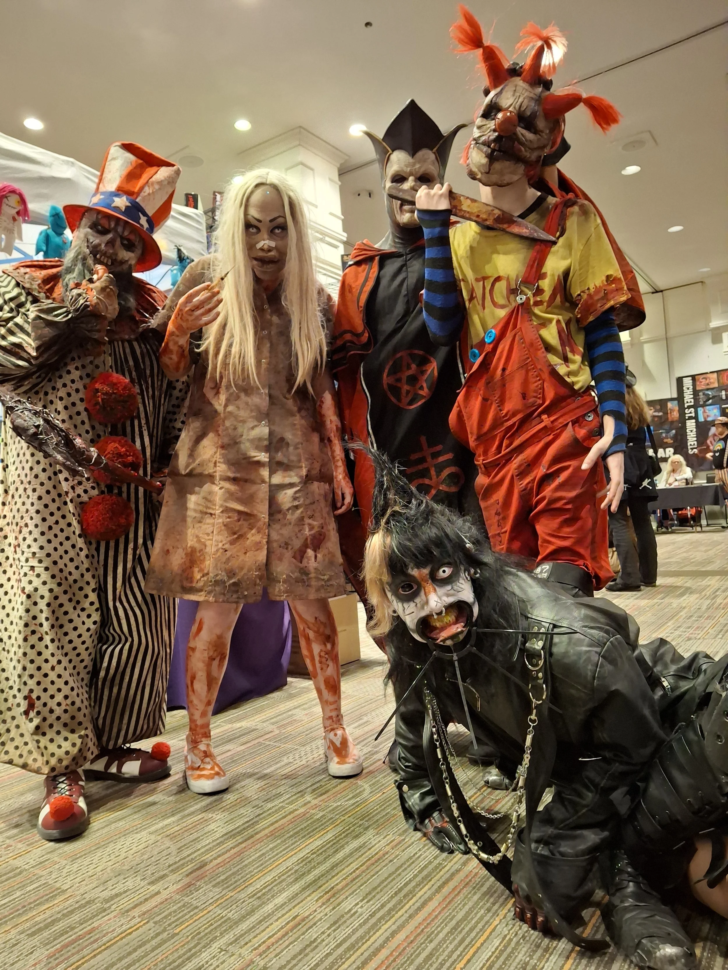 Group of people dressed in elaborate Halloween costumes, including a clown, a person with a pig mask, a person with a red mask and yellow shirt, a person with a demon mask, and a person with a zombie-like face, at a Halloween event.