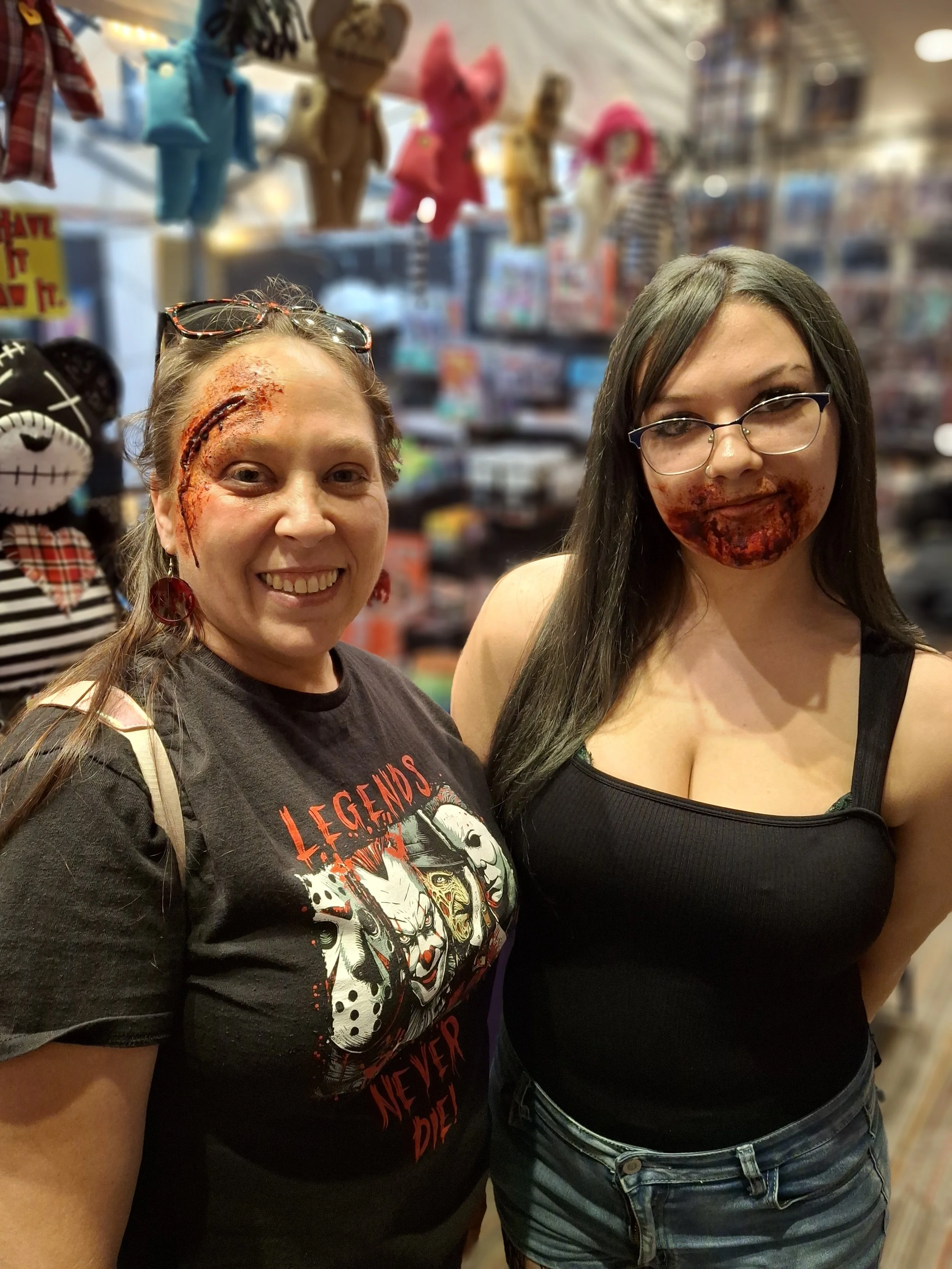 Two women with horror makeup on their faces standing in a toy store, with stuffed animal toys hanging from the ceiling behind them.