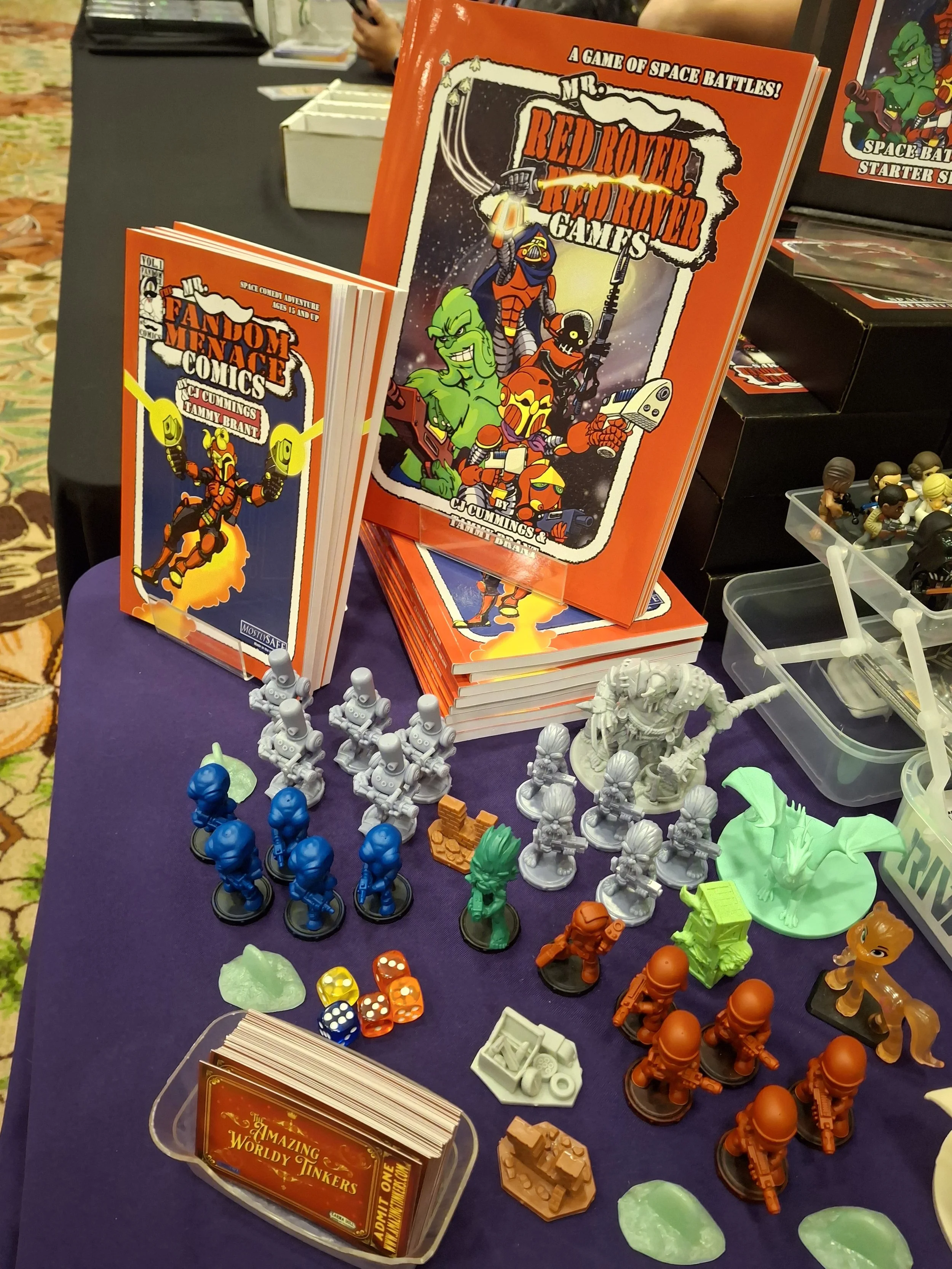 Table display of comic books and miniatures, including titles 'Mr. Fandom Menace Comics' and 'Mr. Red Rover, Red Rover Games,' with various colorful figurines and game materials.