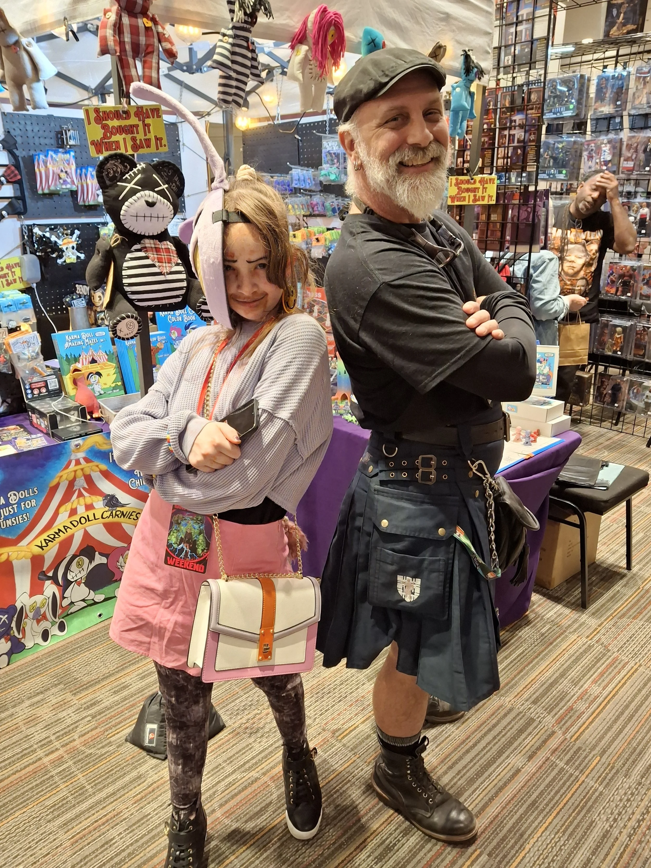 Two people, a young woman and an older man, stand back-to-back at a toy or collectibles store. The young woman is wearing a whimsical headband with plush ears and a long pink tail, a gray sweatshirt, a pink skirt, and patterned tights. The older man 