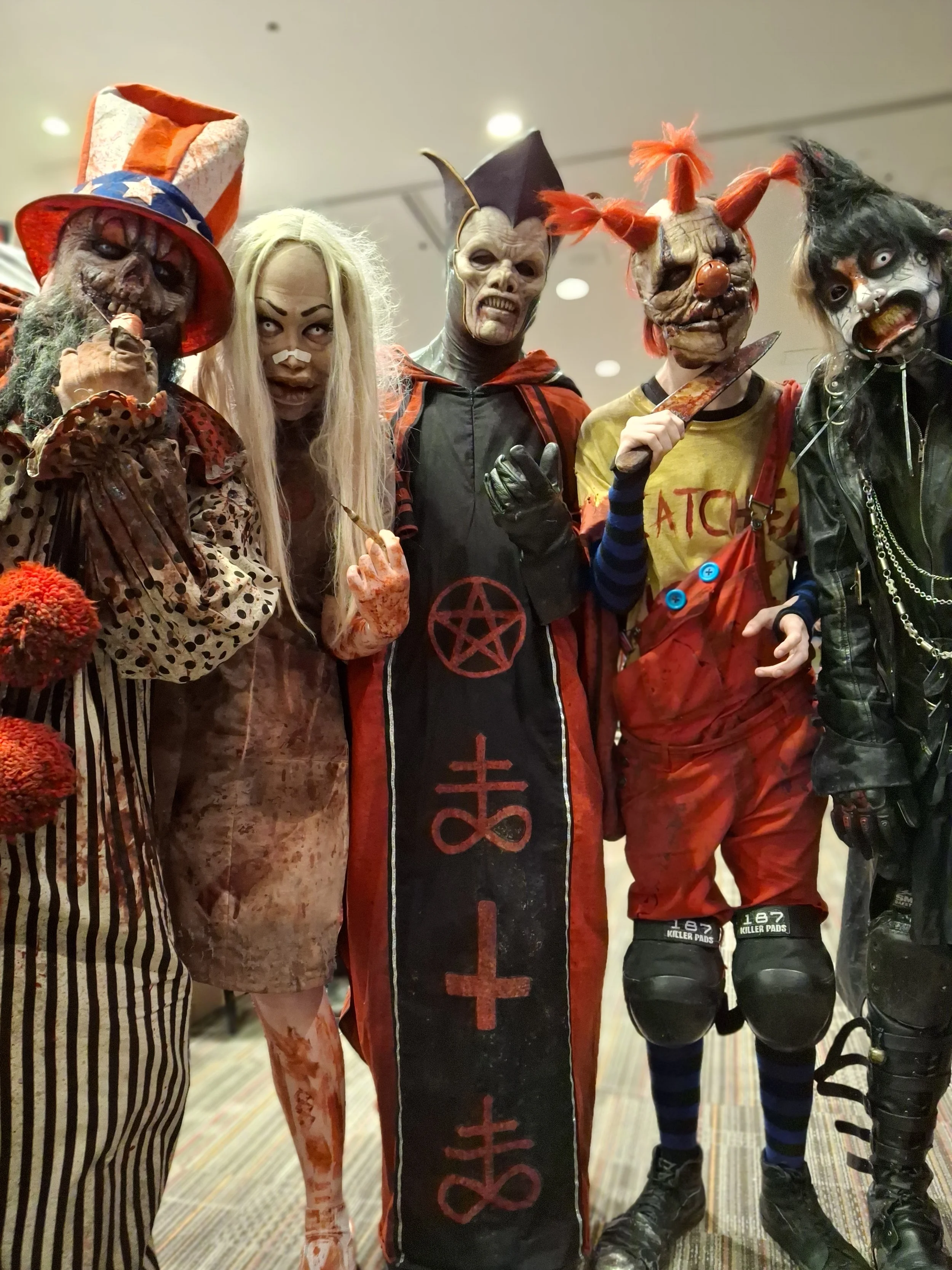 Group of five people dressed in creepy clown and monster costumes for Halloween, standing indoors on carpeted floor.