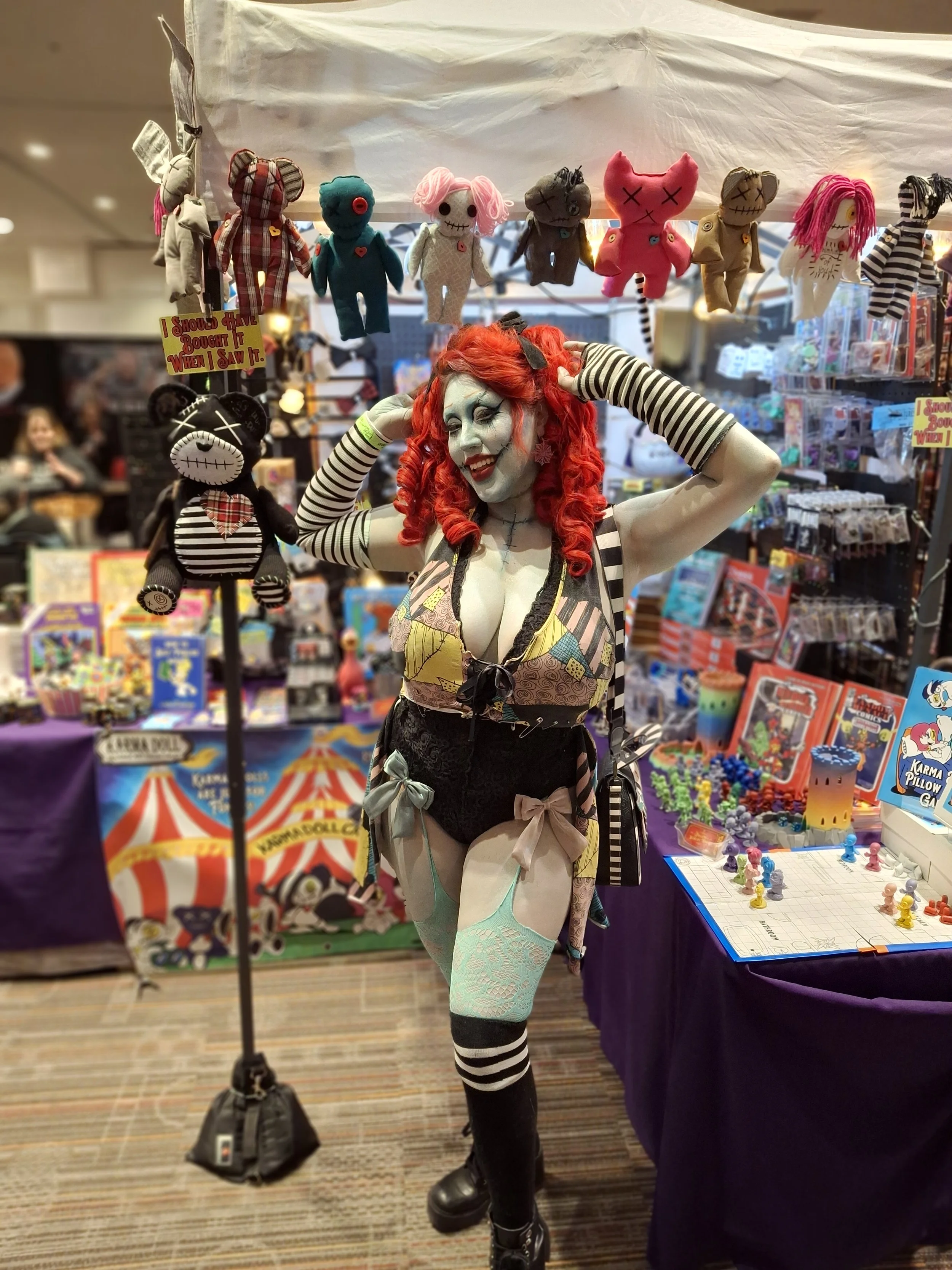 A woman dressed in a theatrical costume with red curly hair, pale makeup, wearing striped long gloves, thigh-high stockings, and boots, posing in front of a display of plush toys and a tabletop with figurines at a convention or pop culture event.