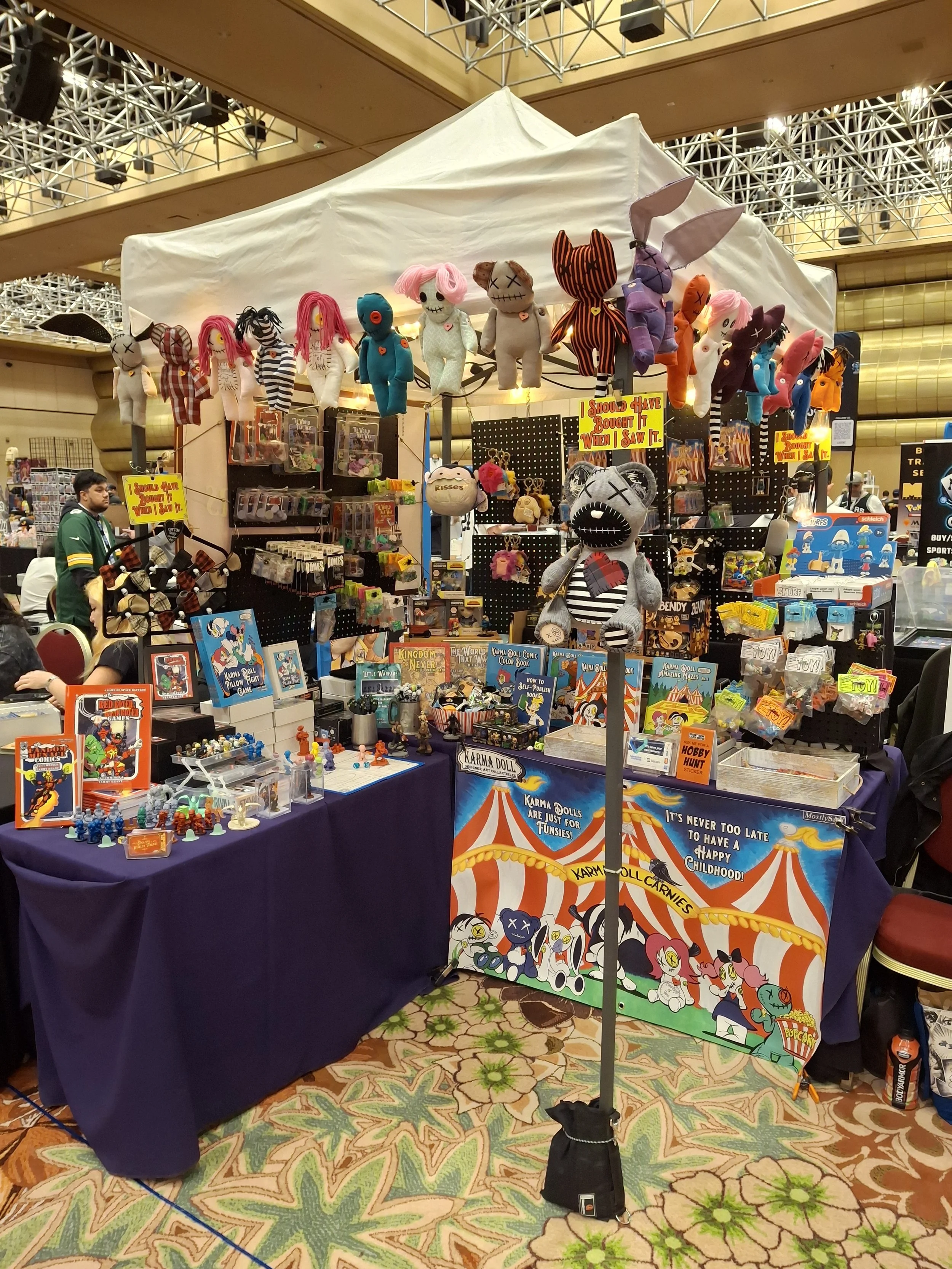 A booth at a convention or fair selling plush toys, toys, and collectibles, with various colorful stuffed animals and merchandise on display, and banners featuring cartoon characters.