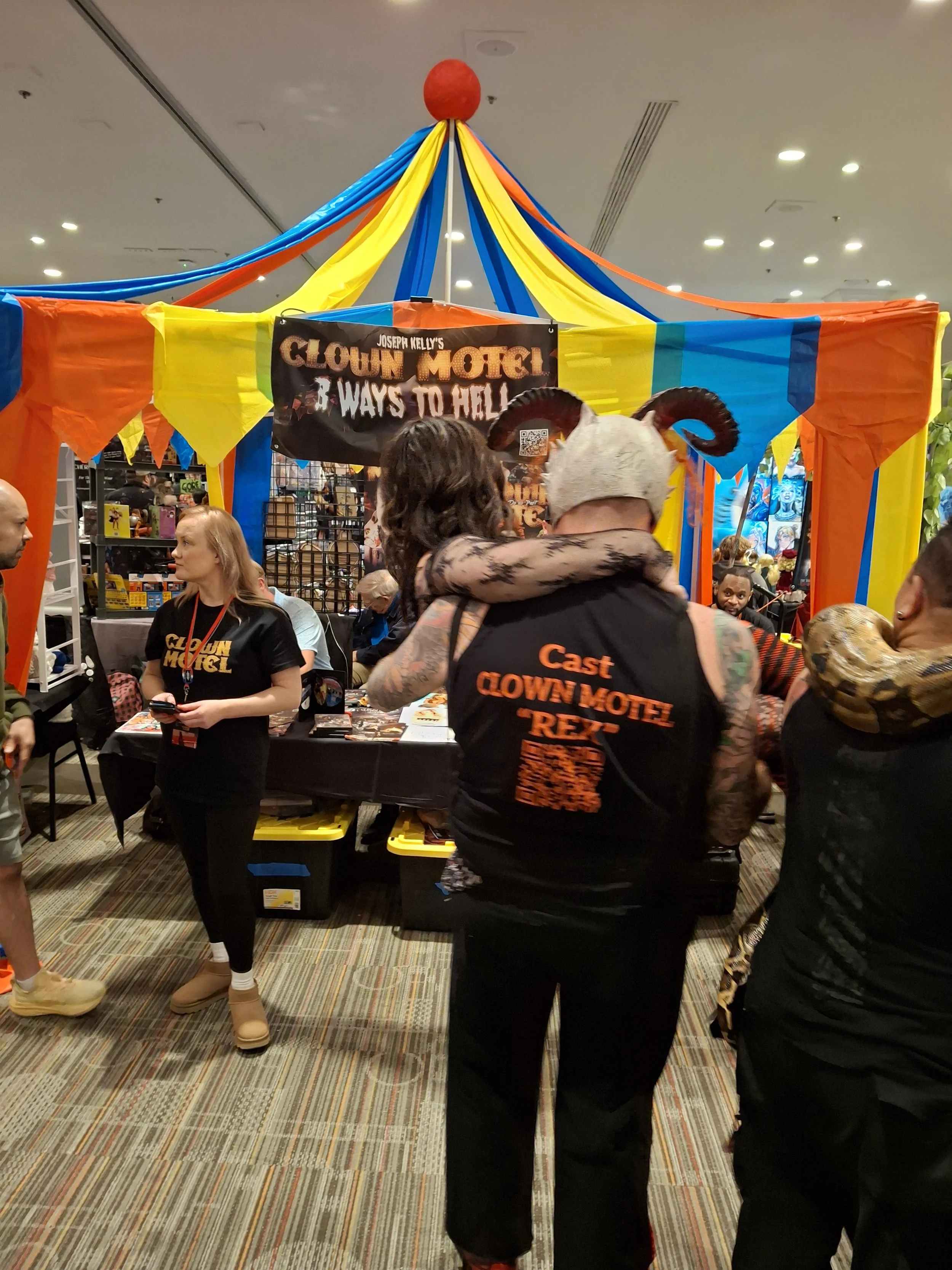 People at a booth promoting Joseph Kelly's Clown Motel, with a colorful tent and a sign listing three ways to hell.