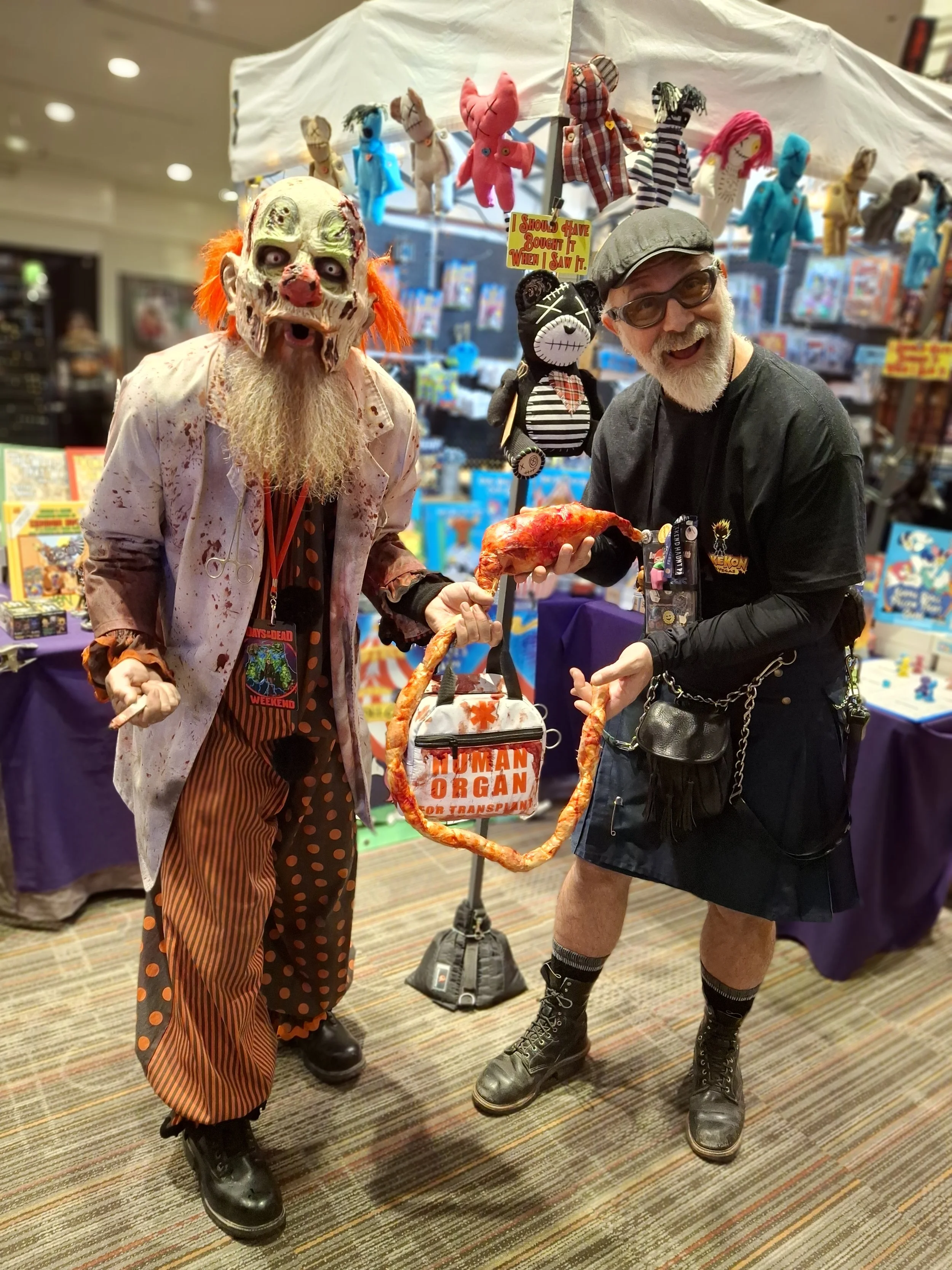 A person in a scary clown costume with a mask and clown makeup, holding a puppet and a fake human organ attached to a ring, standing next to a man in casual attire at a convention booth.