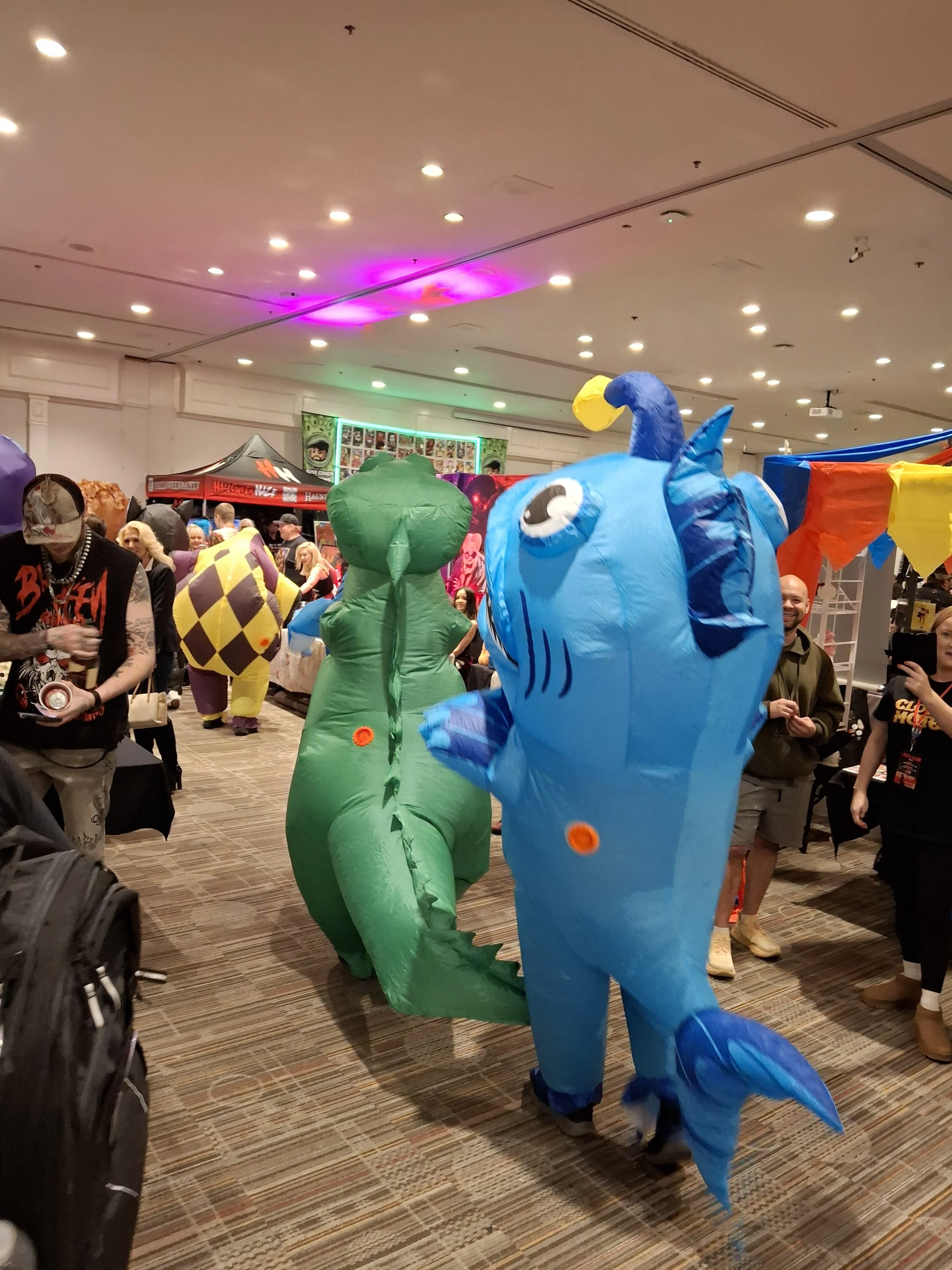 People dressed in colorful mascot costumes including a blue fish and a green dinosaur at a crowded indoor event or convention.
