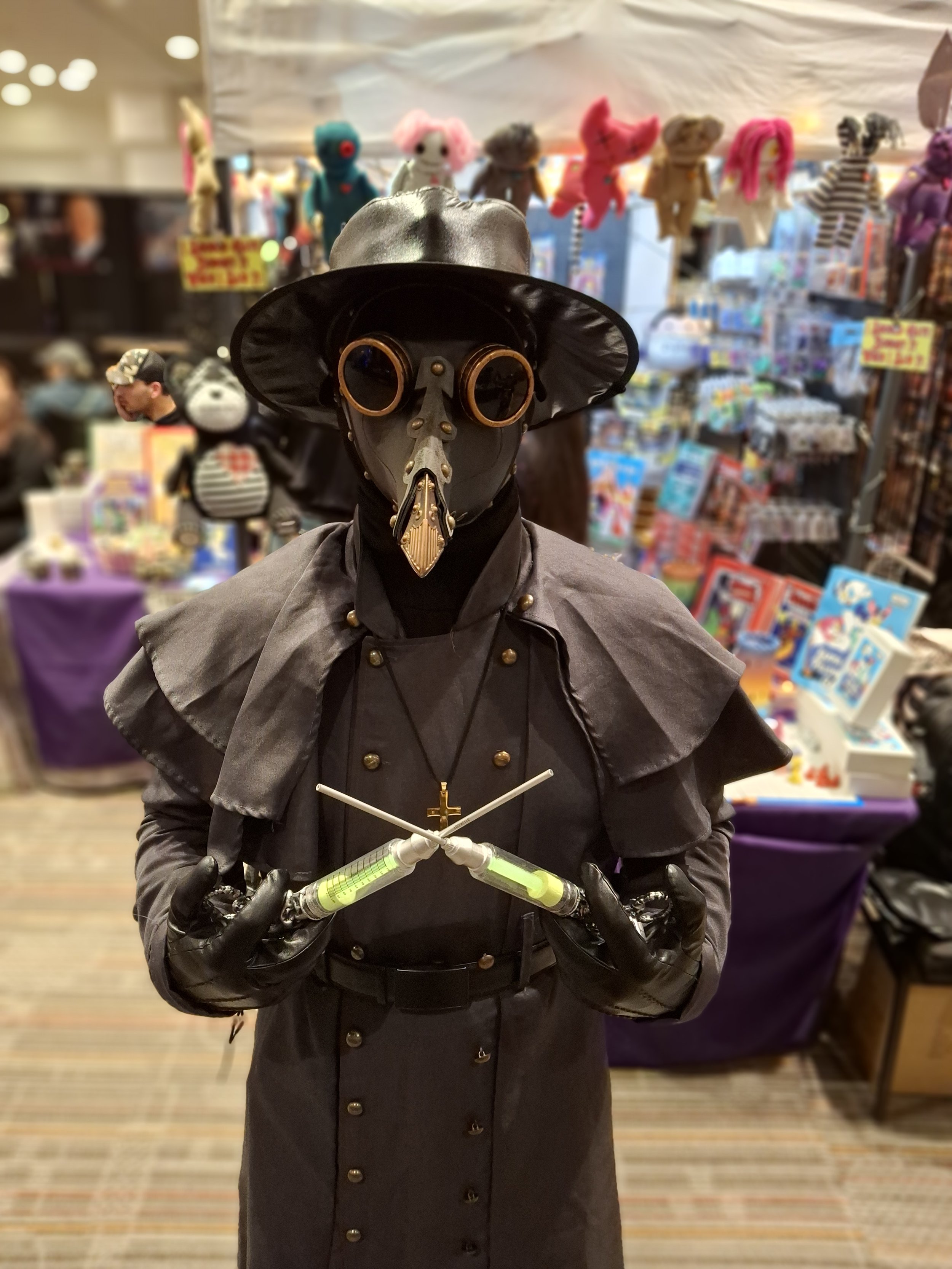 A person dressed as a plague doctor with a beak mask, round goggles, black hat, black coat, and gloves, holding two syringes with green liquid at an indoor event with toys and colorful displays in the background.