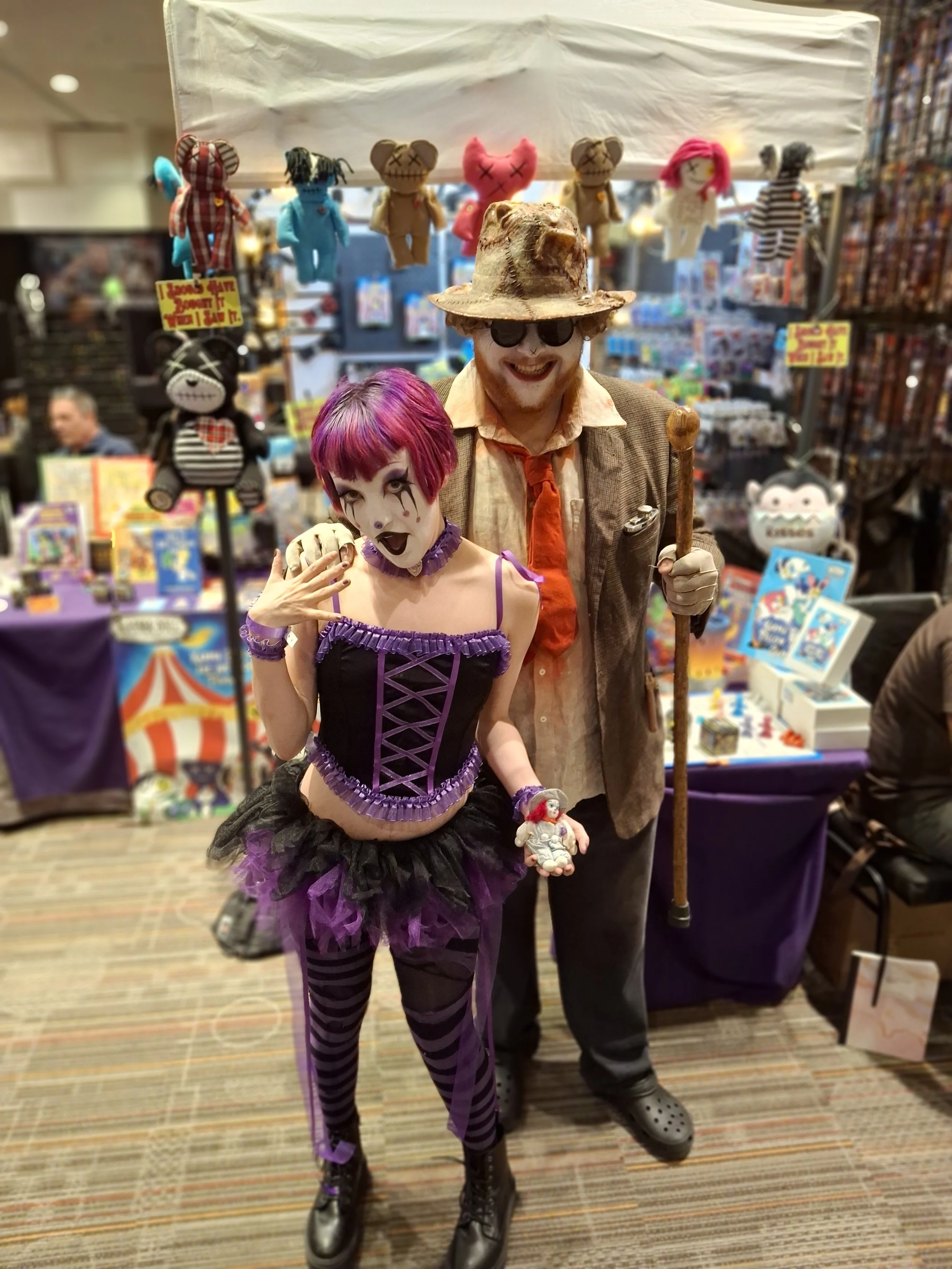 Two people in costumes at a convention, one dressed as a clown with colorful makeup and gothic attire, the other in a teddy bear costume with sunglasses and a hat, standing in front of a booth with plush toys and artwork.