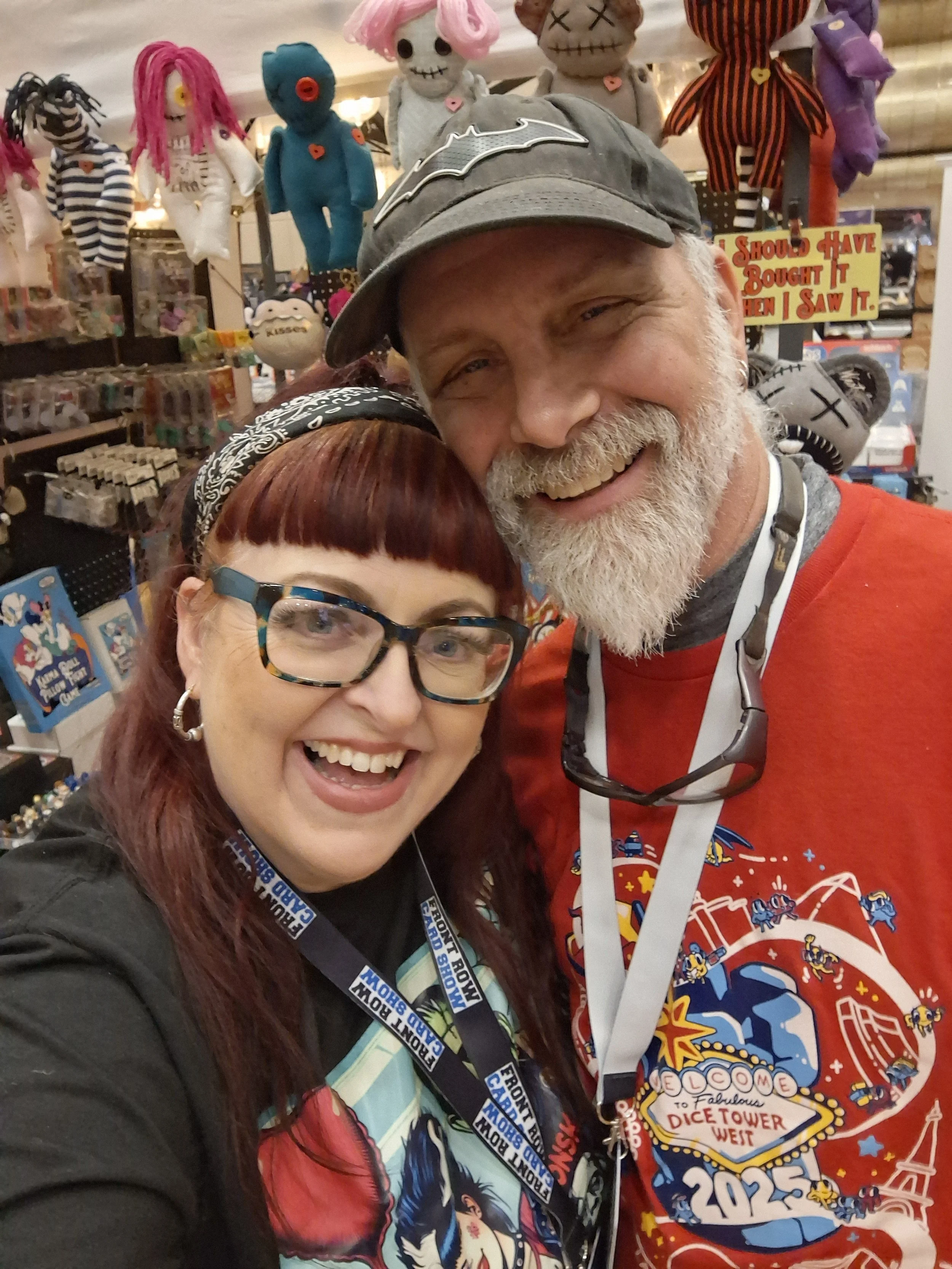 Two people smiling and taking a selfie inside a store with plush dolls, toys, and collectibles in the background. The woman has red hair, glasses, and a black headband, while the man has a gray beard, baseball cap, and a red shirt with a colorful des
