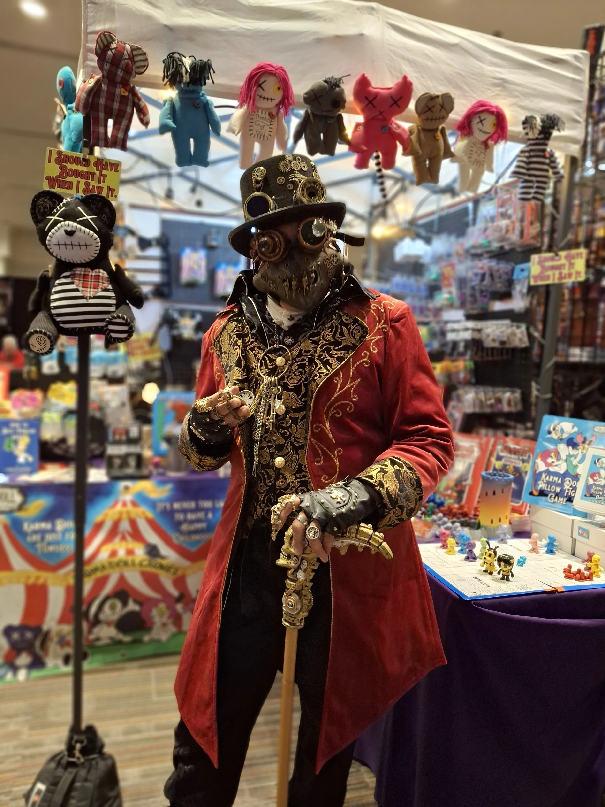 Person dressed in steampunk-inspired outfit with a red velvet coat, decorated with gold patterns, wearing a Victorian style hat with goggles, and jewelry, holding a cane with an ornate handle, surrounded by plush toys and toys on display at a booth.