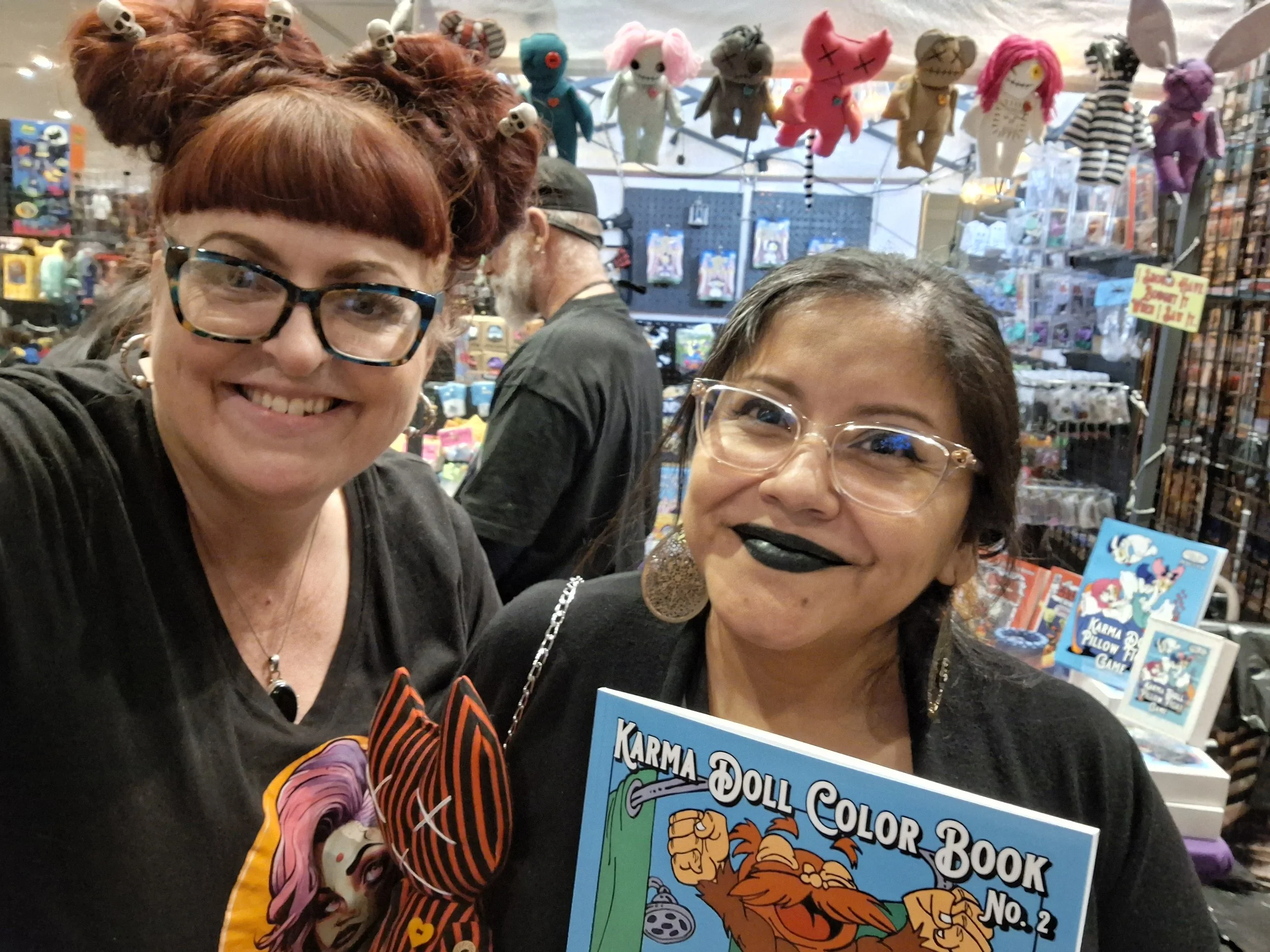 Two women with glasses smiling at the camera inside a store selling plush toys and dolls, with various merchandise and plush characters hanging above in the background.
