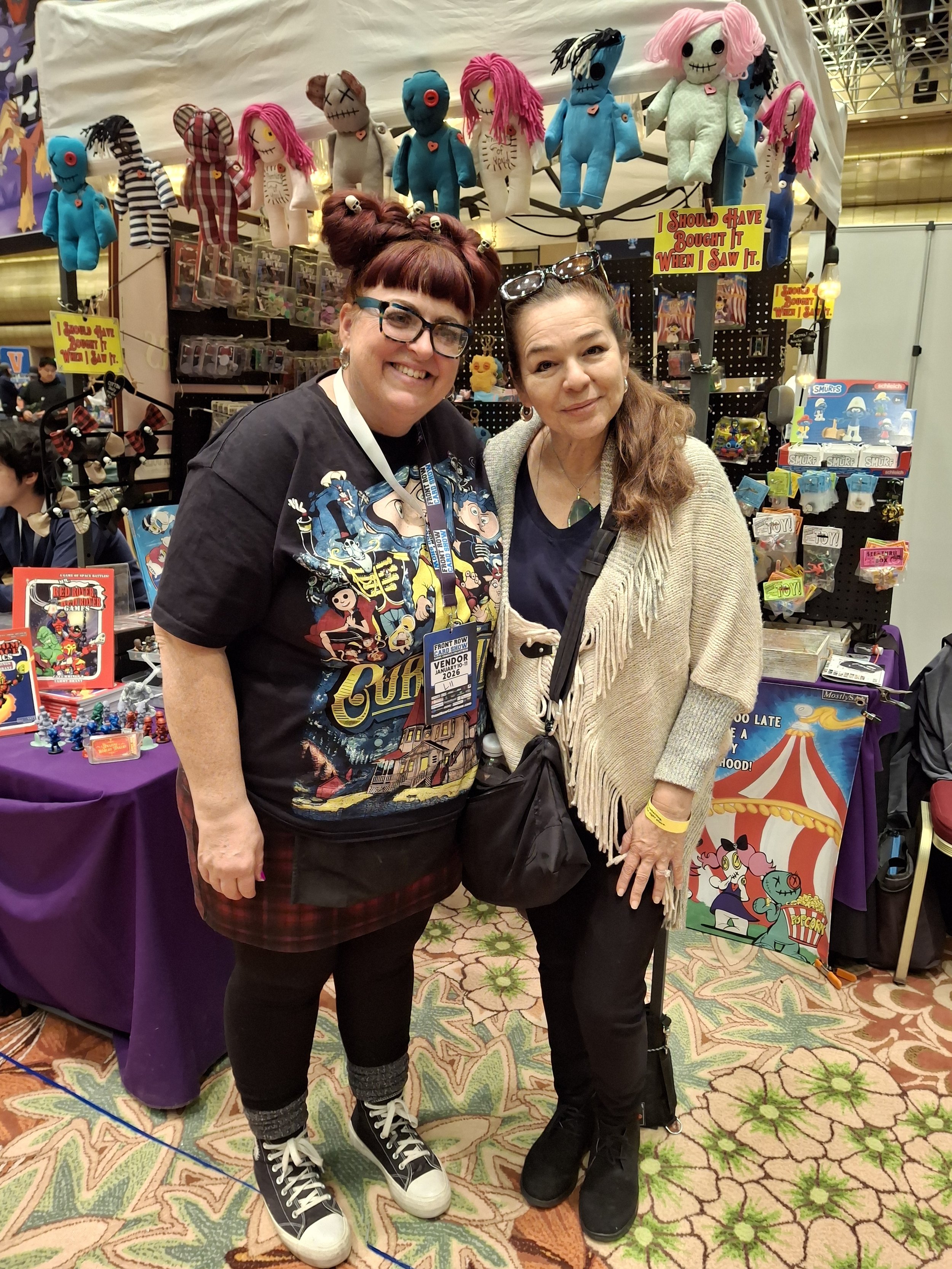 Two women standing together at a convention, smiling. Behind them are toys and plush dolls on display, some with unique designs and colors. The woman on the left is wearing a black T-shirt with colorful graphics, glasses, and sneakers. The woman on t