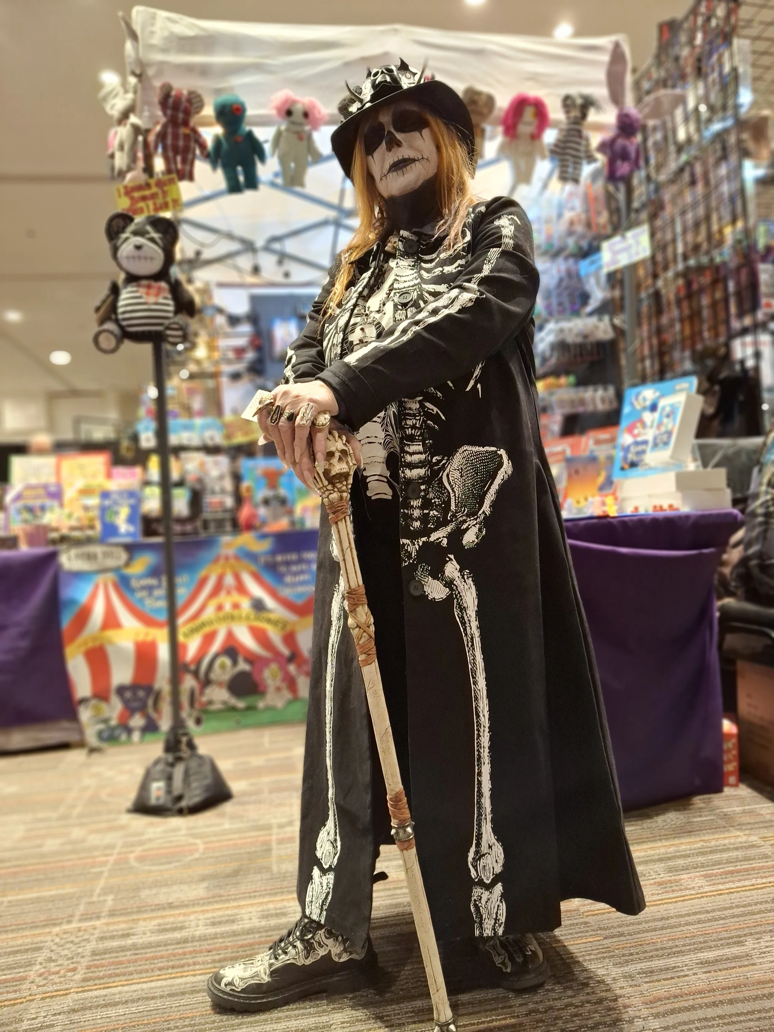Person dressed in skeleton costume with skeleton face paint, wearing a skeleton-themed outfit, holding a skeleton staff, at a toy or collectible store with plush toys and toys in the background.