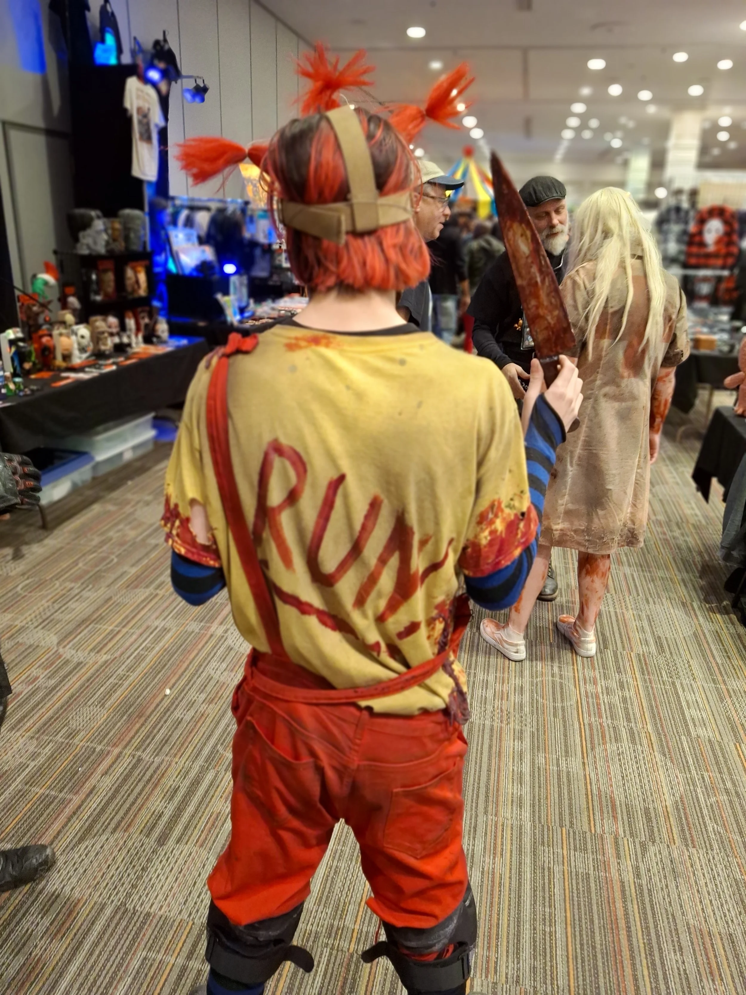 Person in horror costume with orange pigtails, blood-stained shirt reading 'RUN' and red pants, holding a large fake knife at a convention or event.