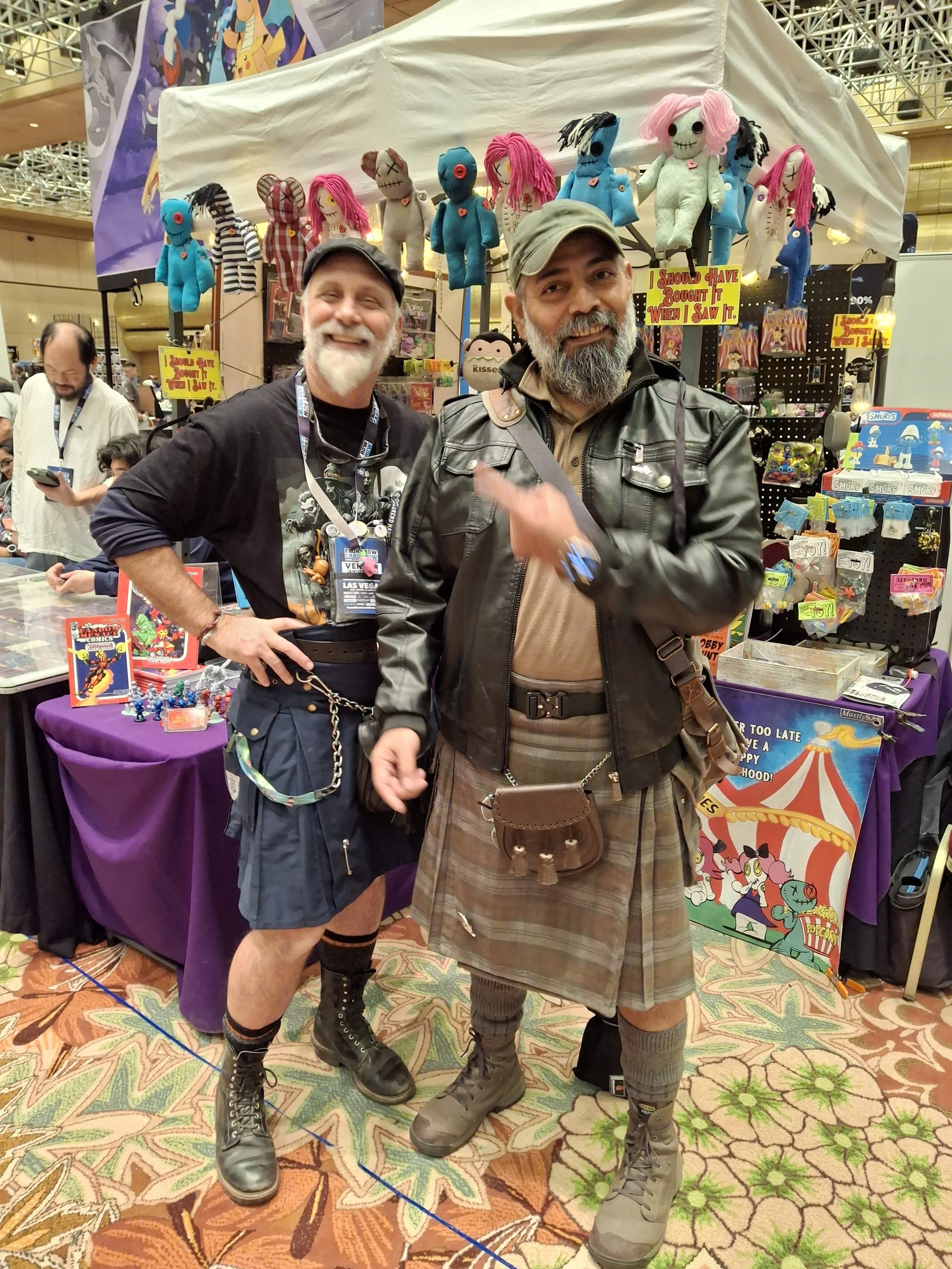 Two men dressed in kilts and leather jackets at a convention, posing for a photo, with plush dolls and other merchandise displayed behind them.