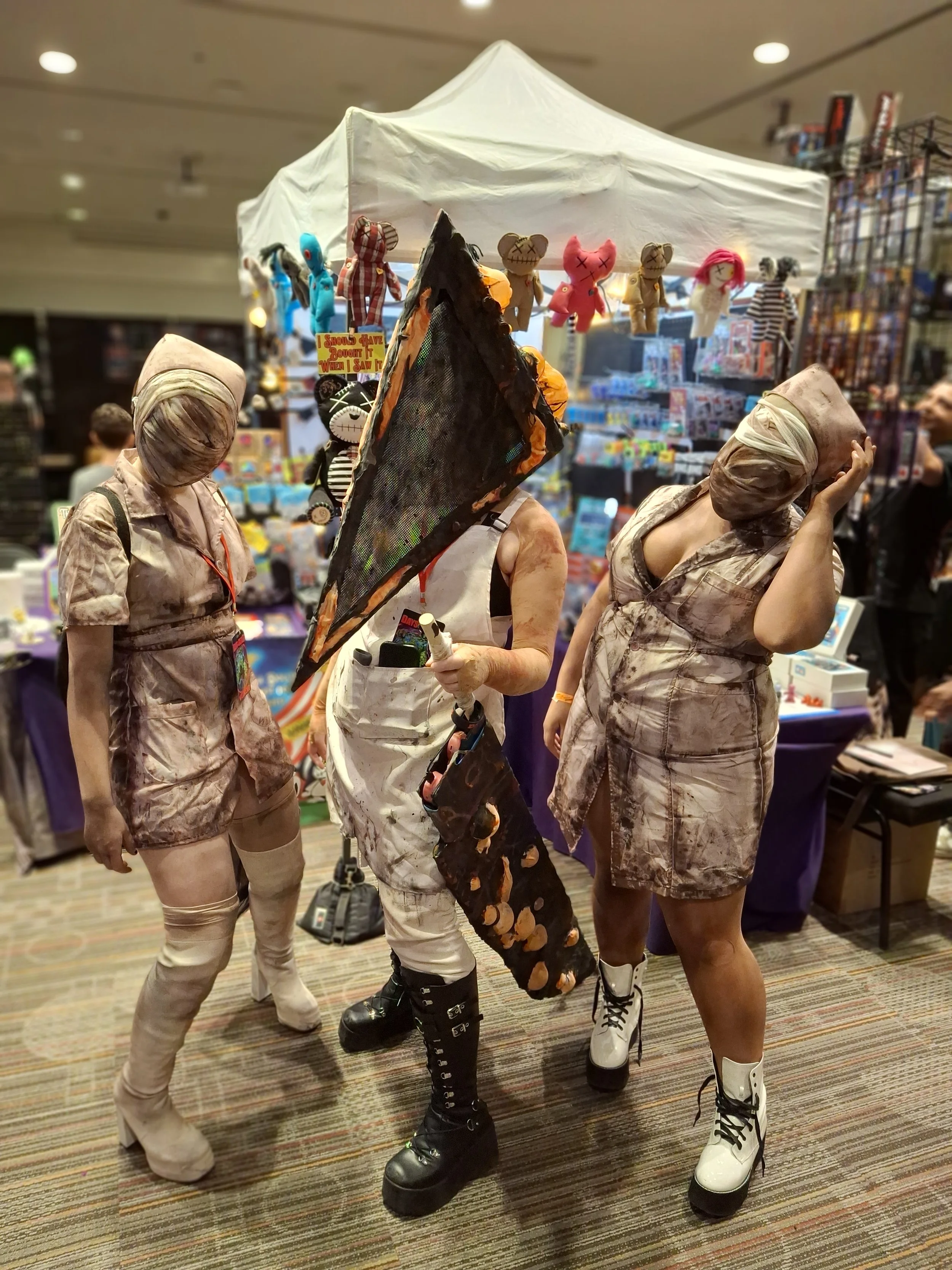 Three people dressed in costumes resembling mummy or zombie characters at a convention or event, with a booth displaying plush toys behind them.