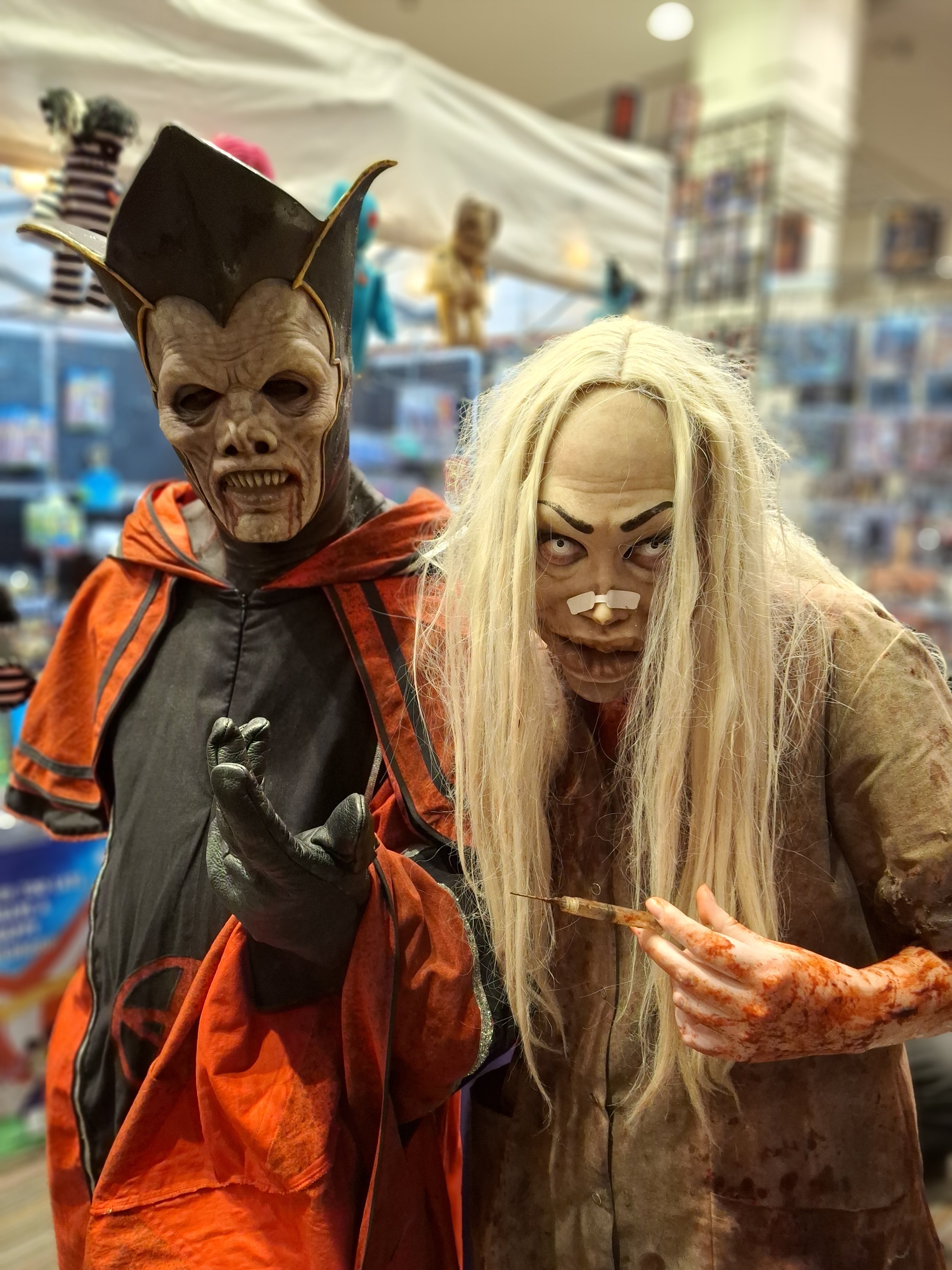 Two people dressed in Halloween costumes, one as a scary demon-like figure with a mask and the other as a bloody nurse with a syringe, in a Halloween store.