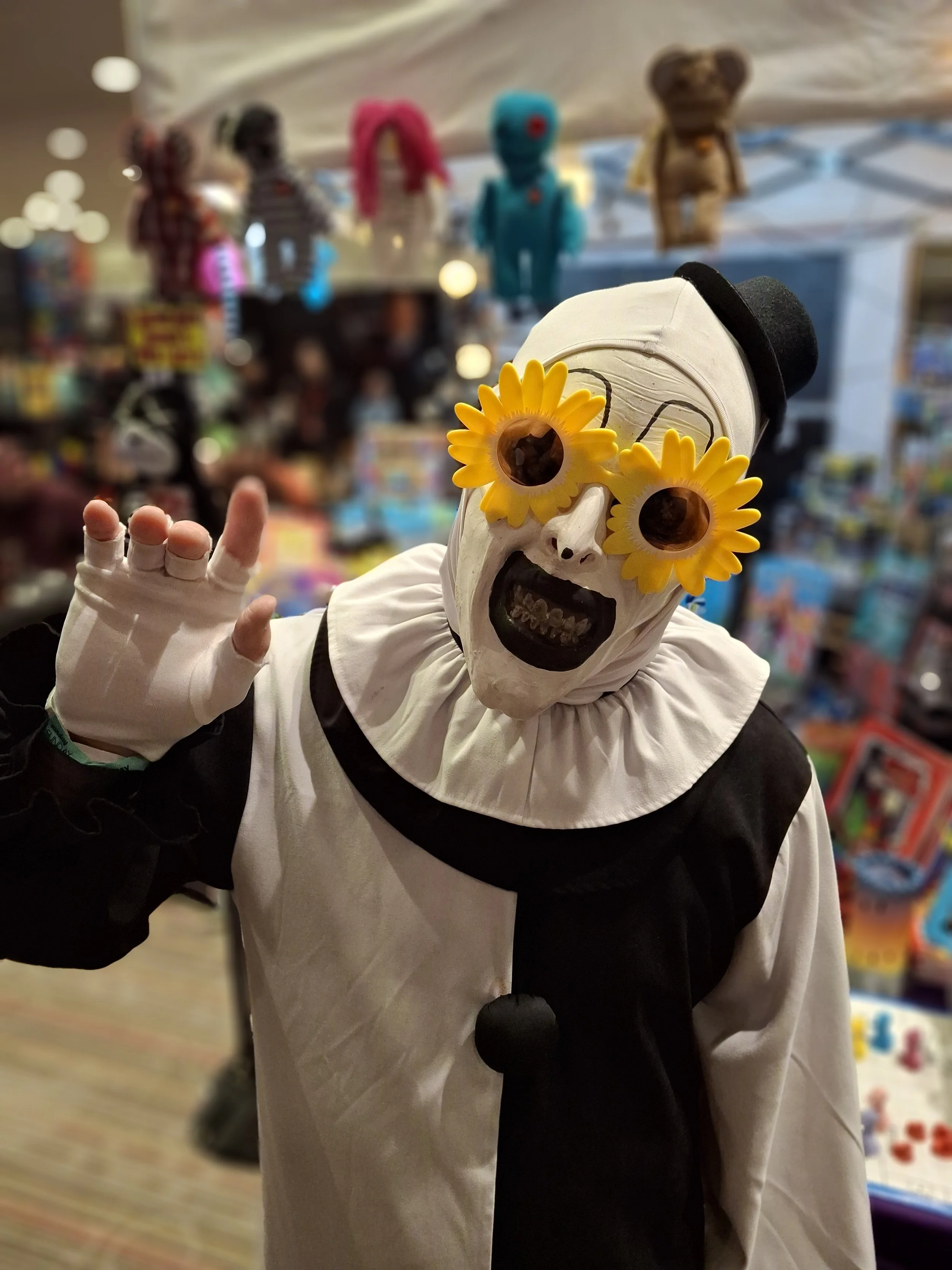 Person dressed as a clown with a skull mask, sunflower sunglasses, wearing a black and white clown costume with a ruffled collar, and a black top hat with a skull emblem, waving in a toy store with colorful plush toys and toys on shelves in the backg