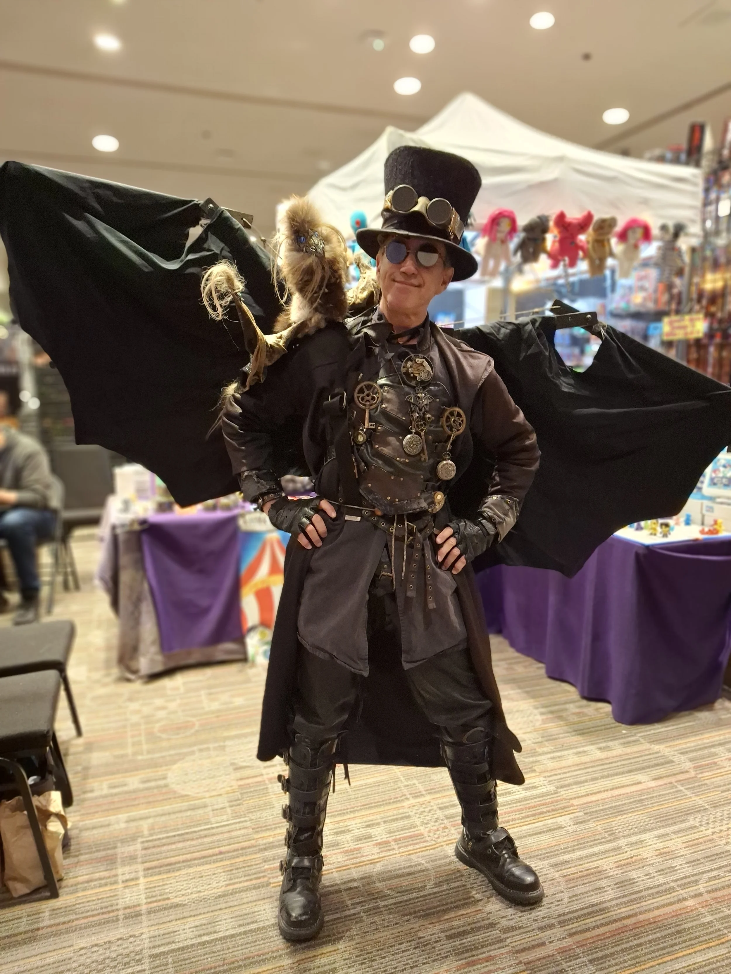 A person dressed in steampunk costume with goggles, gears, and clock pendants, wearing a tall top hat with goggles, and large black bat-like wings. They are standing indoors at a convention or event, with tables and display booths in the background.